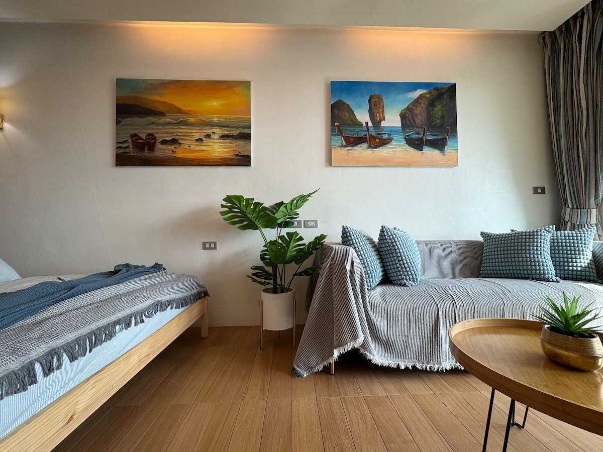 A bright living space features a cozy sofa adorned with patterned cushions and a stylish table. A spacious bed with soft linens is situated nearby, accompanied by two landscape paintings on the walls and a leafy plant adding a touch of greenery.