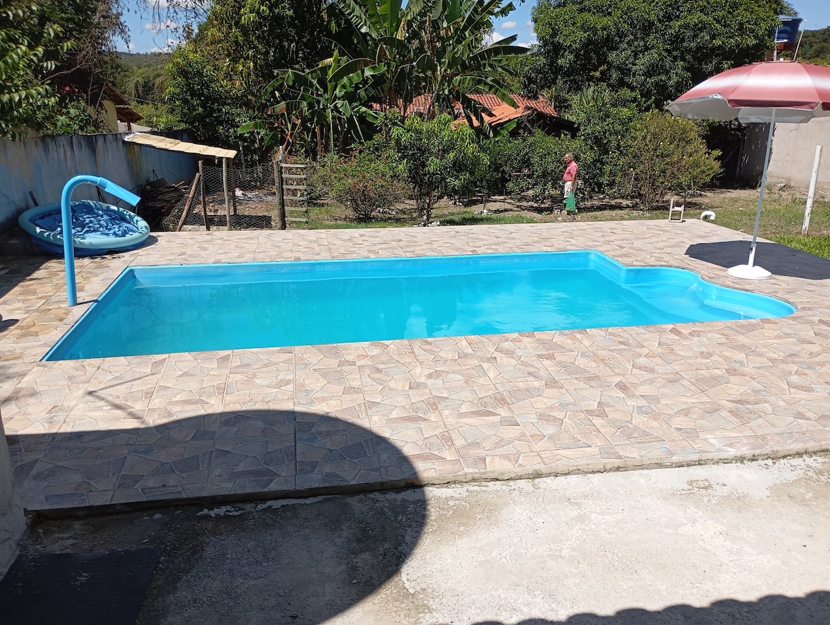 A spacious swimming pool is set within a tiled area, surrounded by greenery and residential structures in the background. Sun loungers are positioned beside the pool, and a shaded area is provided by an umbrella. The overall setting conveys a tranquil outdoor space.