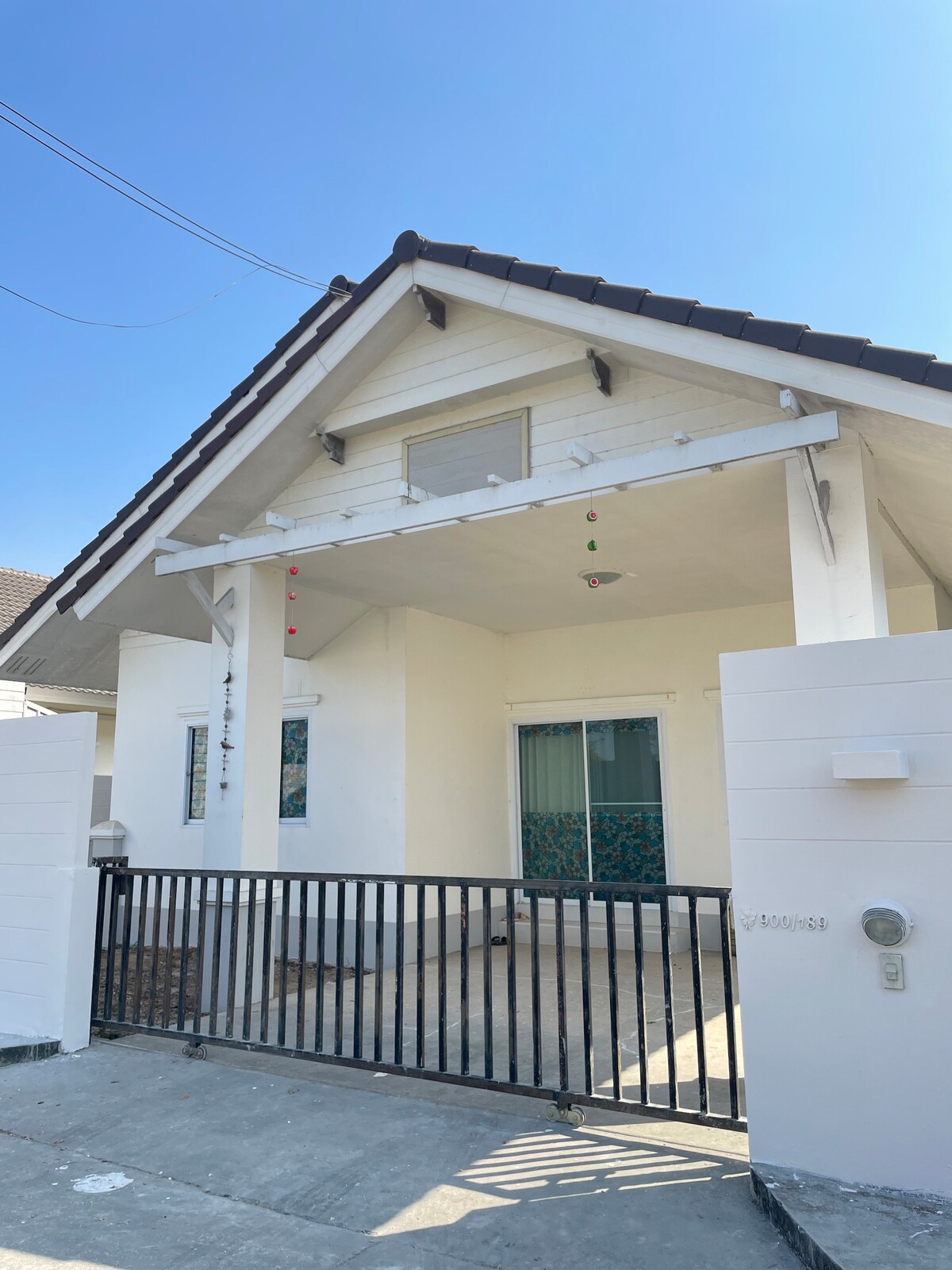 A white house with a gabled roof features a covered porch. Brightly colored decor hangs from the eaves, adding a touch of cheer. The front yard is enclosed by a black gate, leading to a spacious area with large windows allowing natural light to enter.