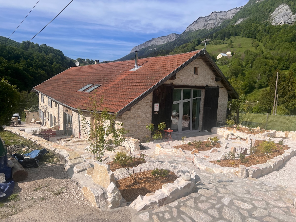 The renovated farmhouse features a charming stone exterior, complemented by a sloping tiled roof. Surrounding gardens are laid out with native plants, while a pathway leads to large glass doors that open to the interior. The landscape showcases green hills and mountains in the background.