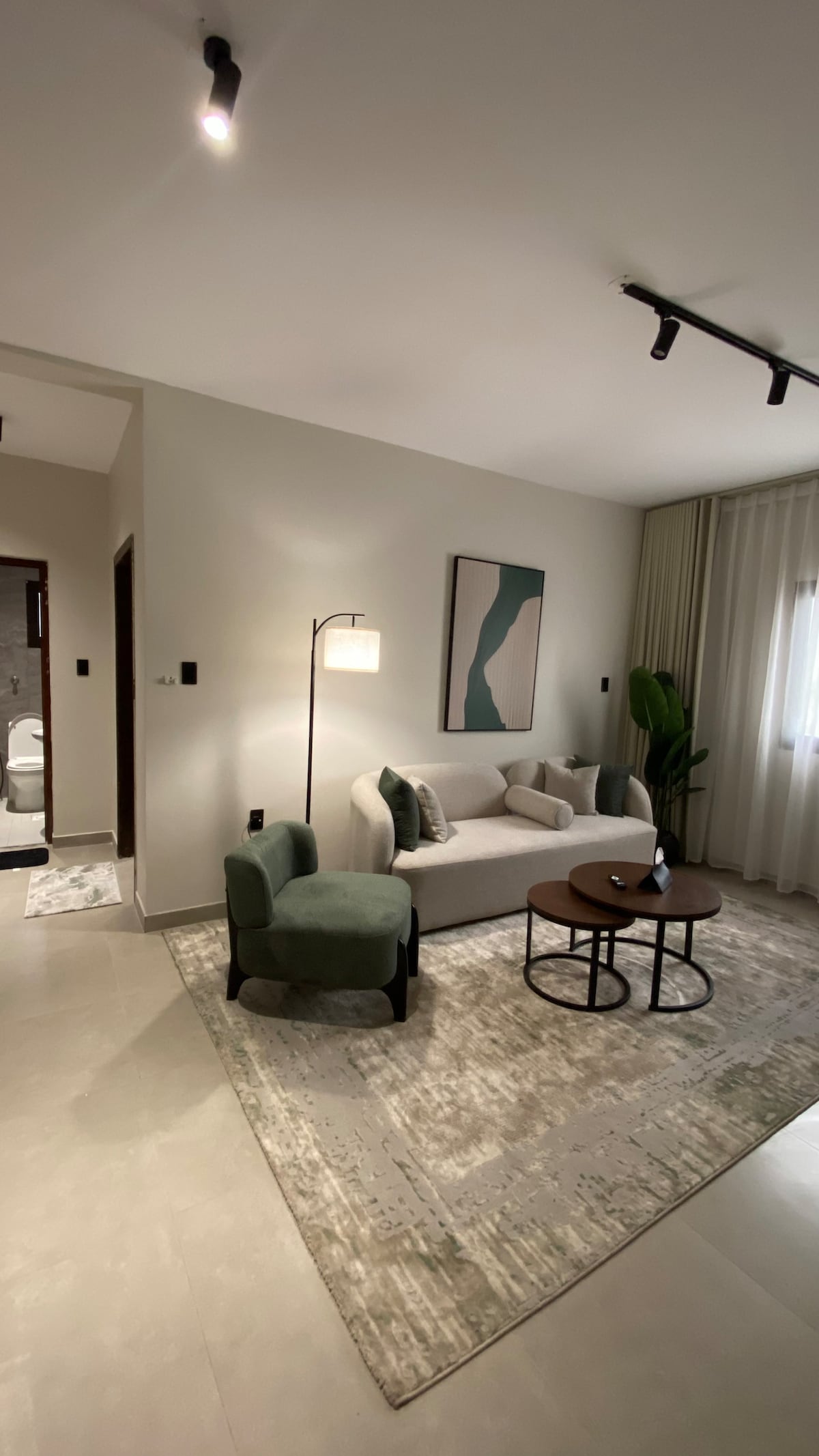 A modern living area features a light-colored sofa paired with green accent chairs. A round coffee table sits at the center, with a small decorative item on it. Soft curtains frame the window, and a floor lamp provides ambient lighting, enhancing the overall layout.