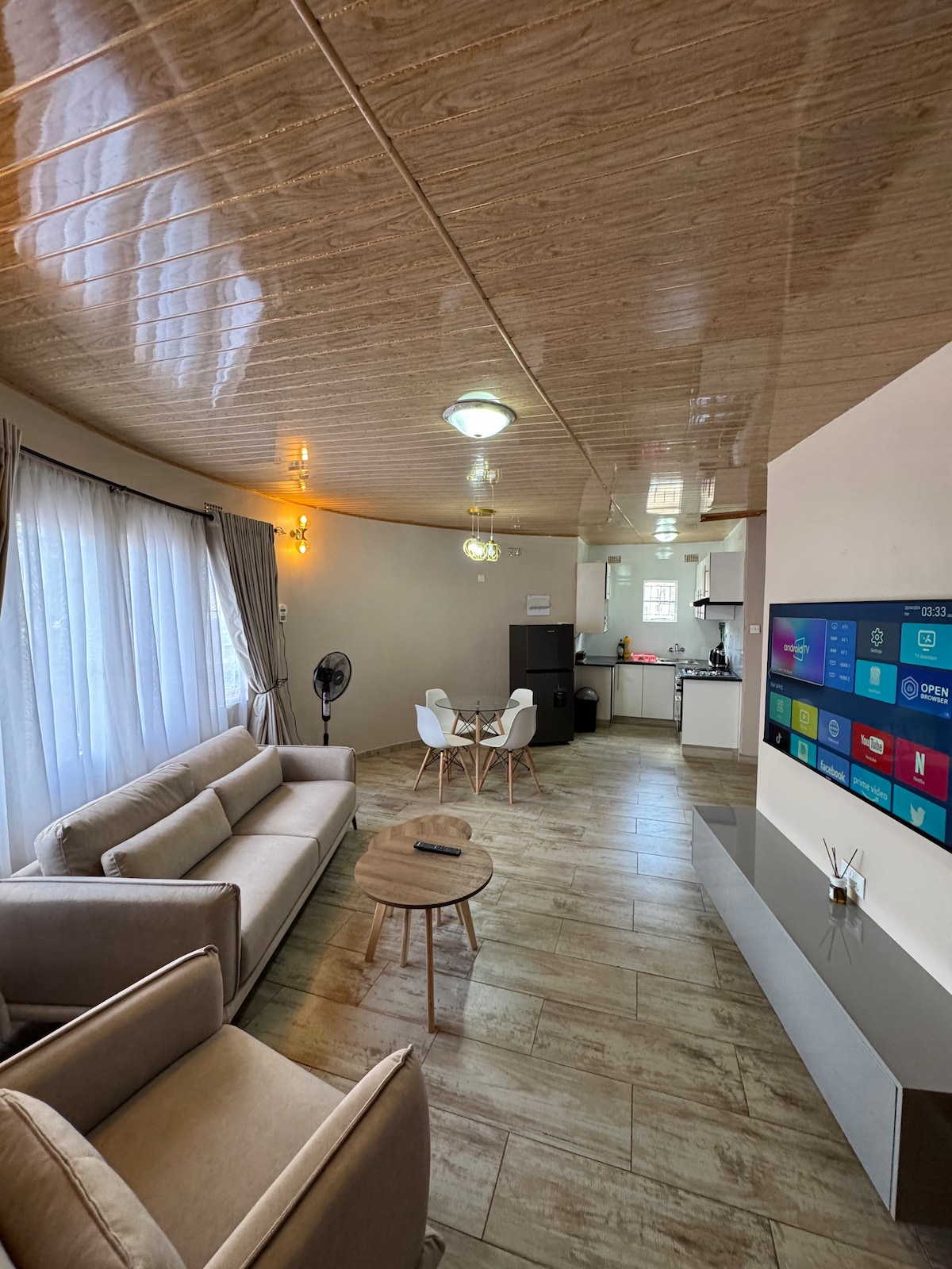 An open-concept living area is presented, featuring a light-colored sofa and two white chairs. A round coffee table sits in the center. Natural light filters through sheer curtains, reflecting off a smooth ceiling. A wall-mounted television displays various app icons, while a kitchen area is visible in the background.