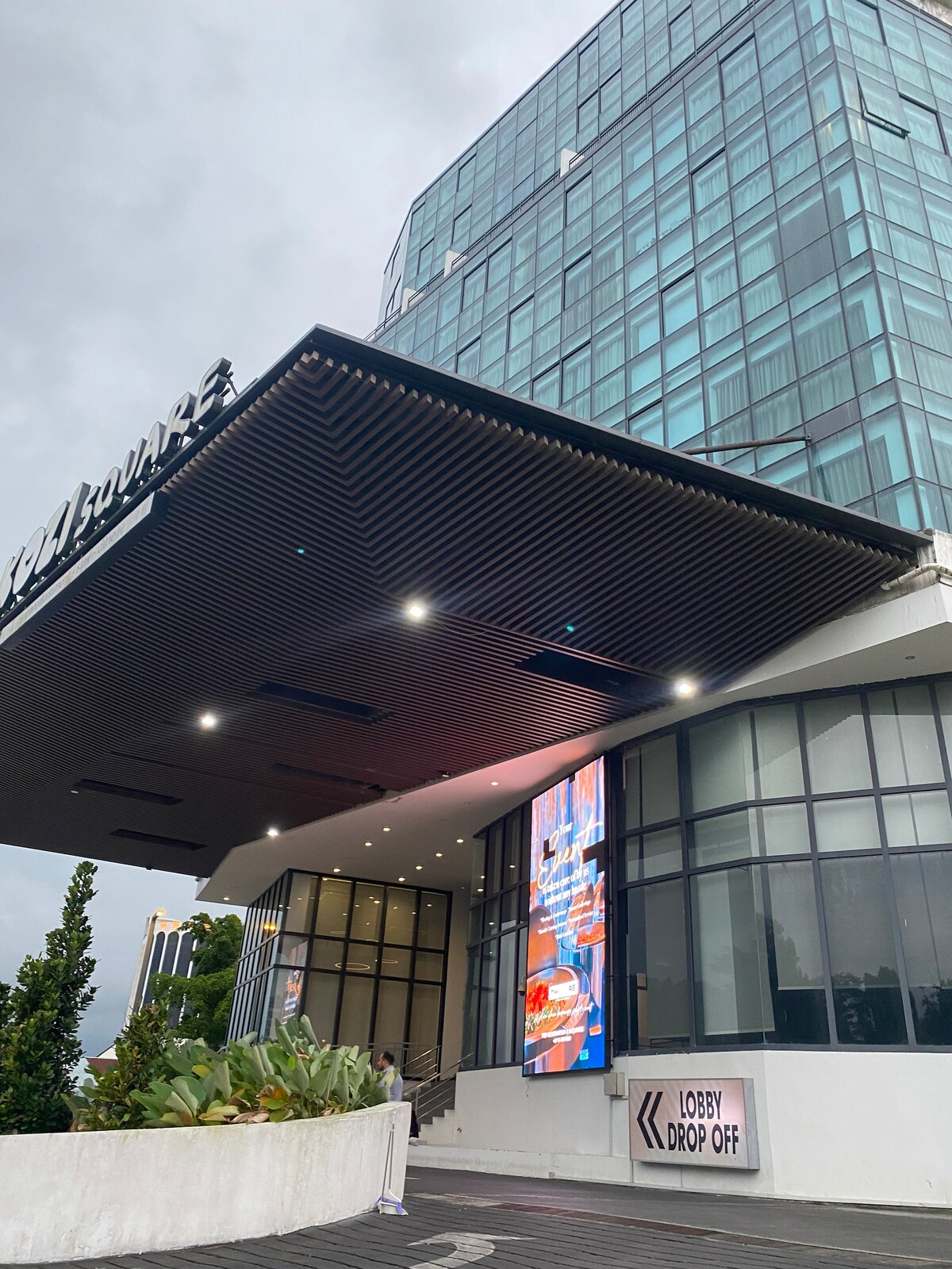 The image features a modern building with a glass facade. A covered entrance is highlighted by bright lights, and signage indicates the lobby drop-off area. Lush greenery is visible in the foreground, adding a touch of nature to the urban setting.