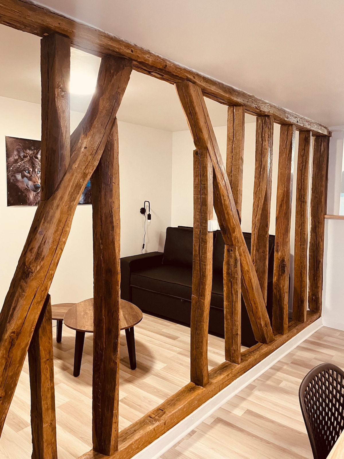 A section of the apartment is framed by rustic wooden beams, adding character to the space. A black sofa is visible alongside a small round table. The clean lines of the light wooden flooring complement the natural tones of the beams.
