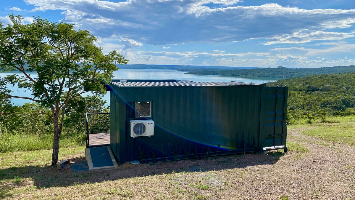 A green shipping container structure is positioned on a grassy hillside, overlooking a tranquil lake. It features a small entrance with a door and a nearby air conditioning unit, framed by a single tree. The sky is partly cloudy, enhancing the natural scenery.