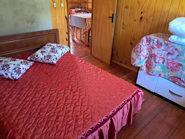 Foto de la cama con una colcha de aluminio rojo y una almohada con estampados de flores rojas.  En la pared opuesta al fotógrafo, se puede ver parte de una cómoda con mantas encima.