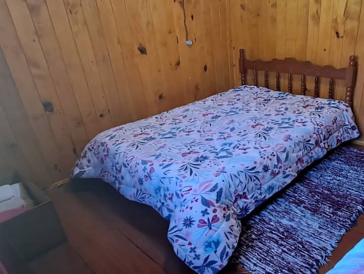 Foto de una cama doble con un edredón floral de mariposas y flores en tonos fríos. La cama está junto a una pared por un lado y a una alfombra que se extiende desde la cabecera hasta el pie de la cama por el otro. En la esquina izquierda hay una cesta con ropa de cama.