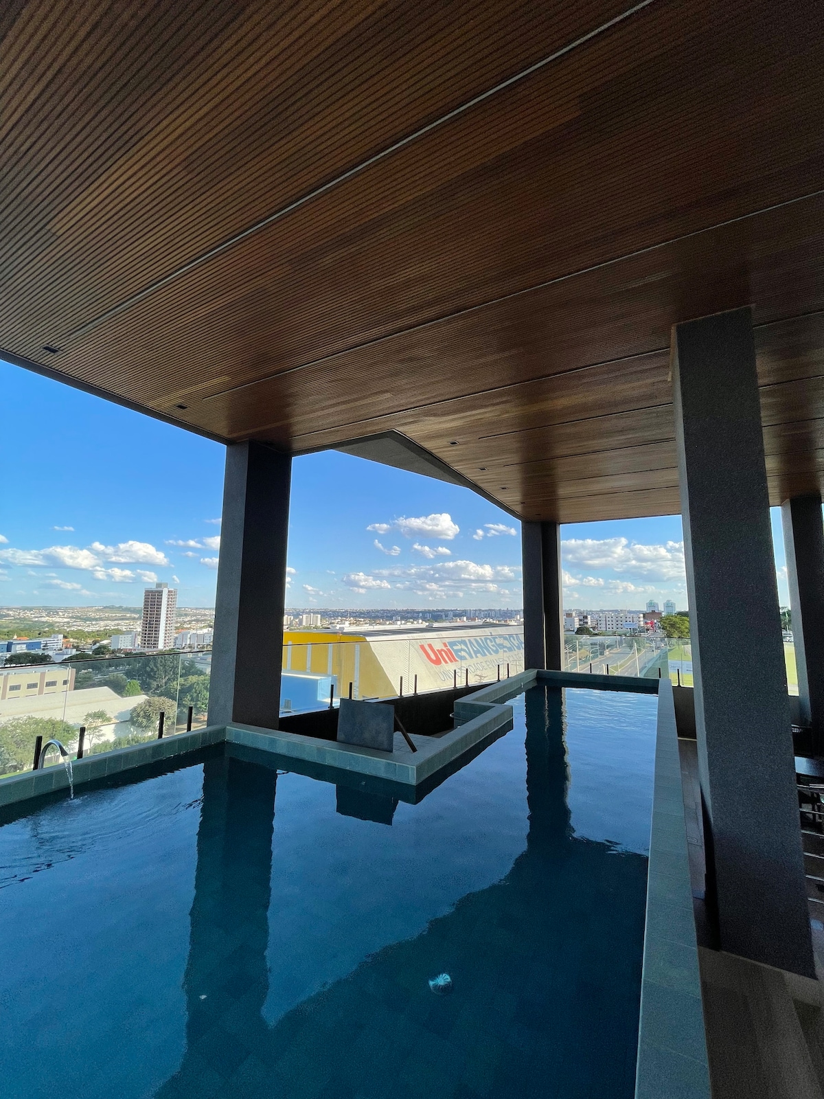 A rooftop pool area features a tranquil water surface reflecting the sky, surrounded by modern architectural elements. Open spaces are created by the expansive wooden ceiling, while clear glass railings provide unobstructed views of the cityscape and distant clouds.