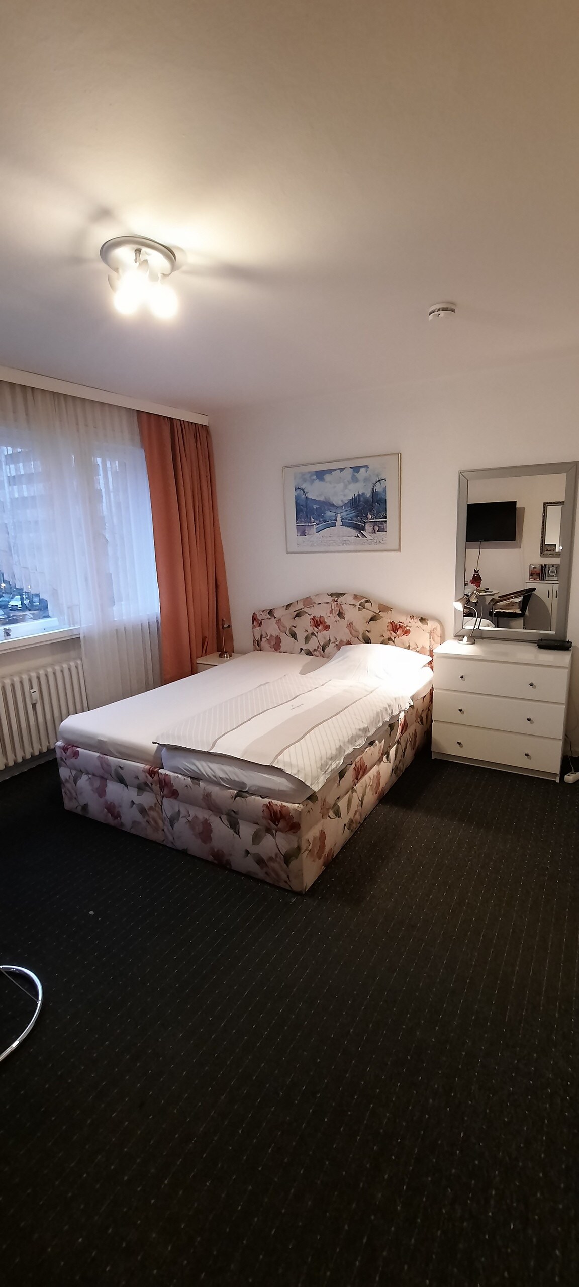 A well-lit bedroom features a floral-patterned bed with a neatly arranged white duvet. A mirror and a dresser sit adjacent to the bed. Natural light enters through draped curtains, adding to the cozy ambiance of the room.