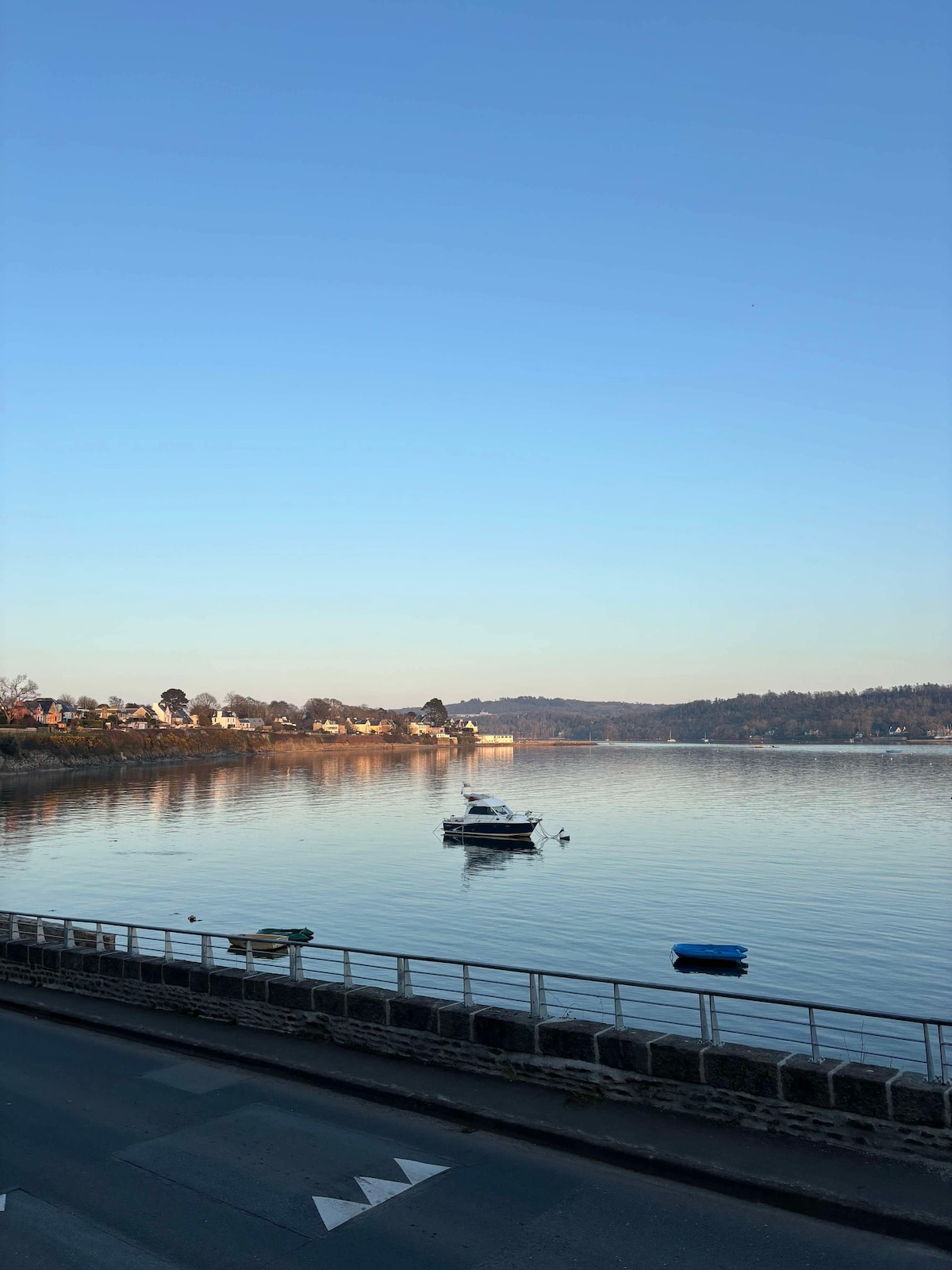 The serene water of the Elorn is captured, reflecting the clear blue sky. A small boat floats gently on the surface, while nearby structures are lined along the shore, with trees and hills visible in the distance.