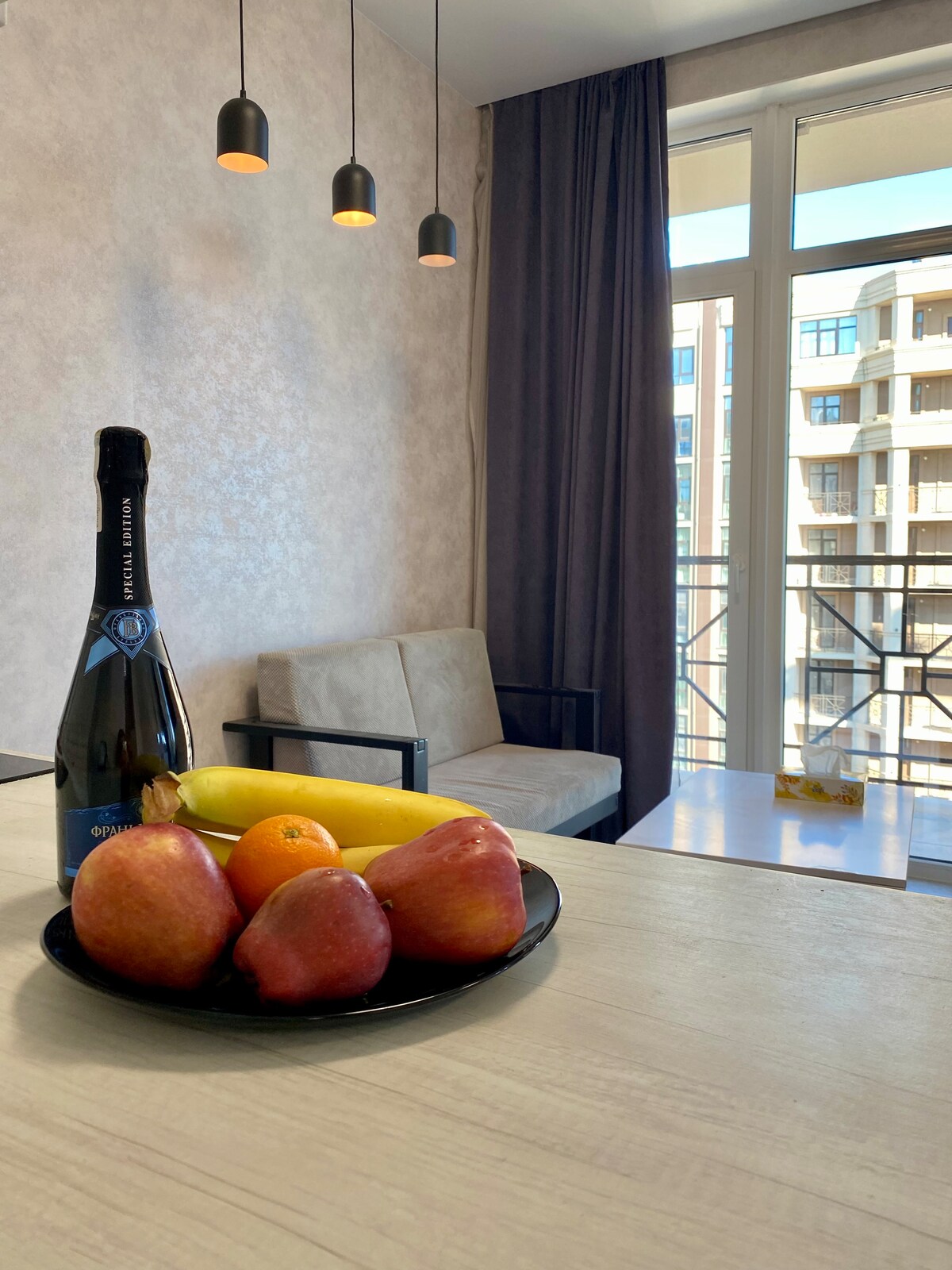 A plate of assorted fruits, including apples, a banana, and an orange, is placed on a light-colored table. In the background, a comfortable sofa and large windows with sheer curtains allow natural light to fill the space.