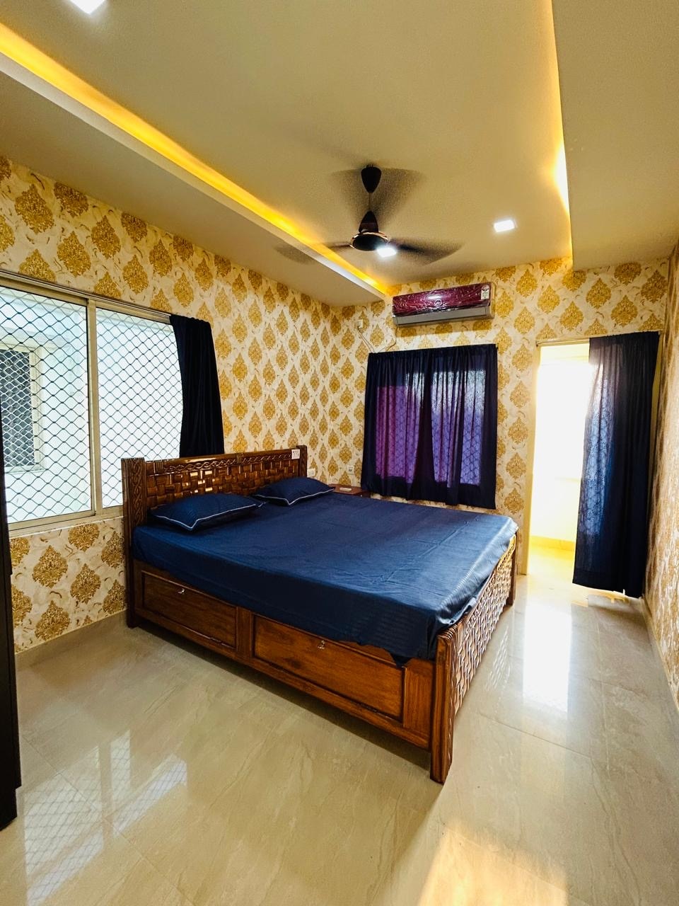 A well-furnished bedroom features a spacious bed with a wooden frame and a blue bedding set. Large windows are fitted with mesh screens and dark curtains, allowing natural light to enter. A door leads to an adjoining area, enhancing the overall spaciousness.