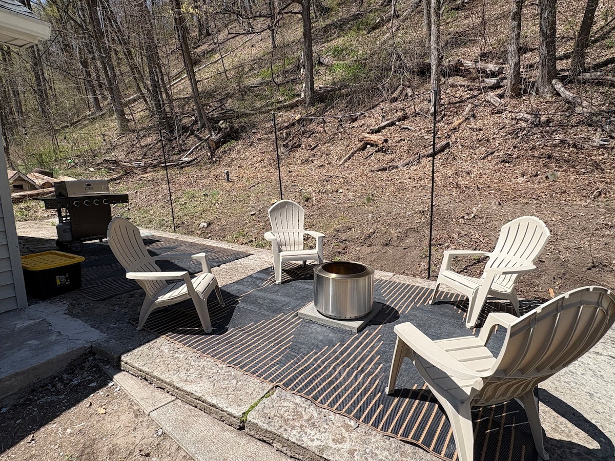 An outdoor fire pit area is set up with four light-colored Adirondack chairs arranged around a circular metal fire pit. A barbecue grill is visible nearby, with a natural backdrop of trees and a sloped landscape in the distance.
