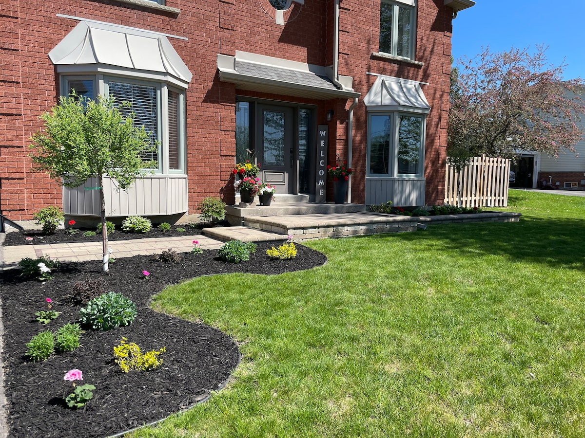 The entrance of the property features a well-maintained landscape with flower beds showcasing colorful blooms and a variety of plants. A brick exterior is complemented by large windows, and a front porch is framed by decorative awnings, creating an inviting and charming entryway.