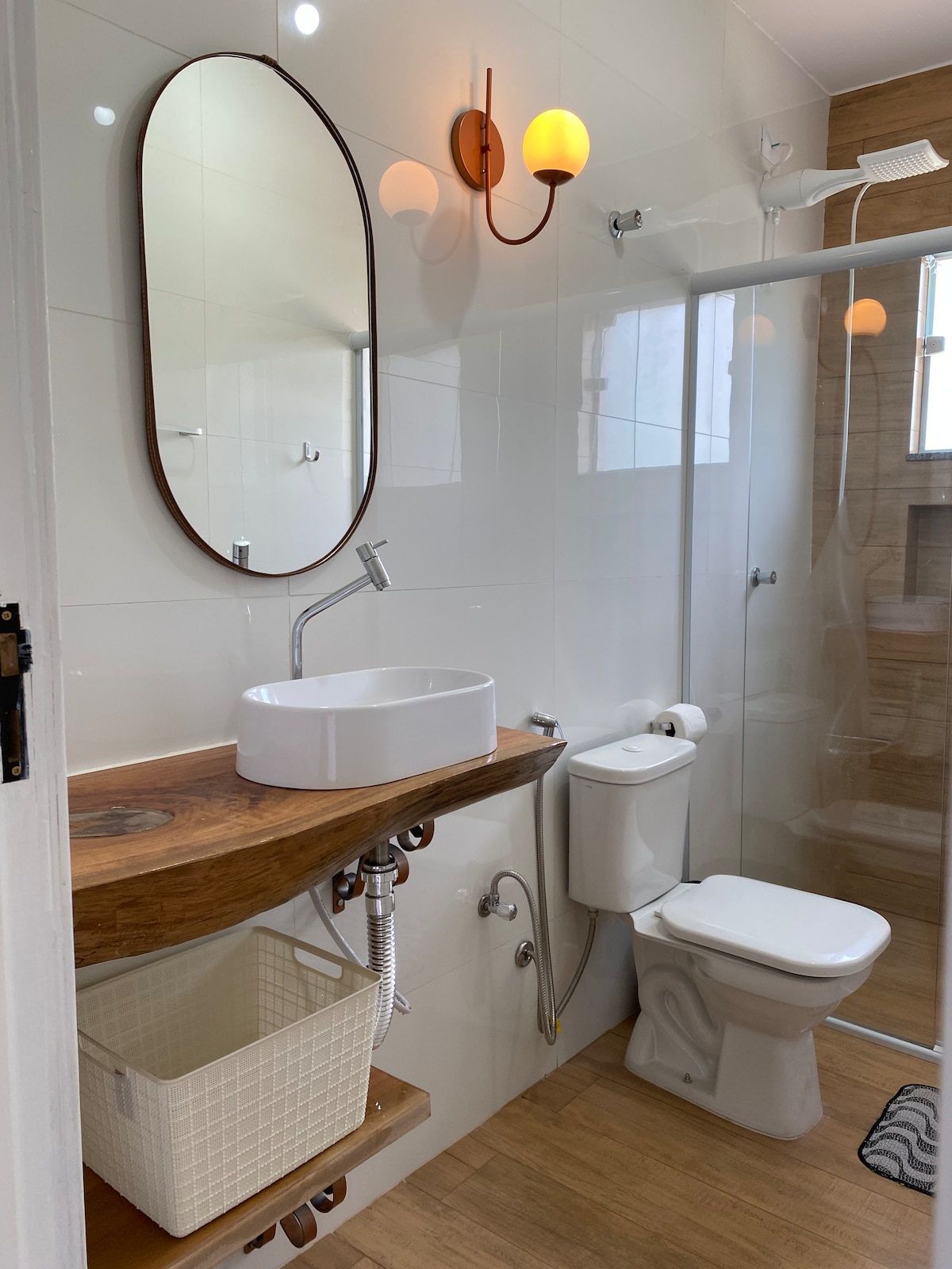 A modern bathroom features a wooden shelf with a round sink and a stylish wall-mounted mirror. A clear glass shower enclosure is seen, with a showerhead positioned above. A toilet is situated beside the shower, complemented by natural light from a nearby window.