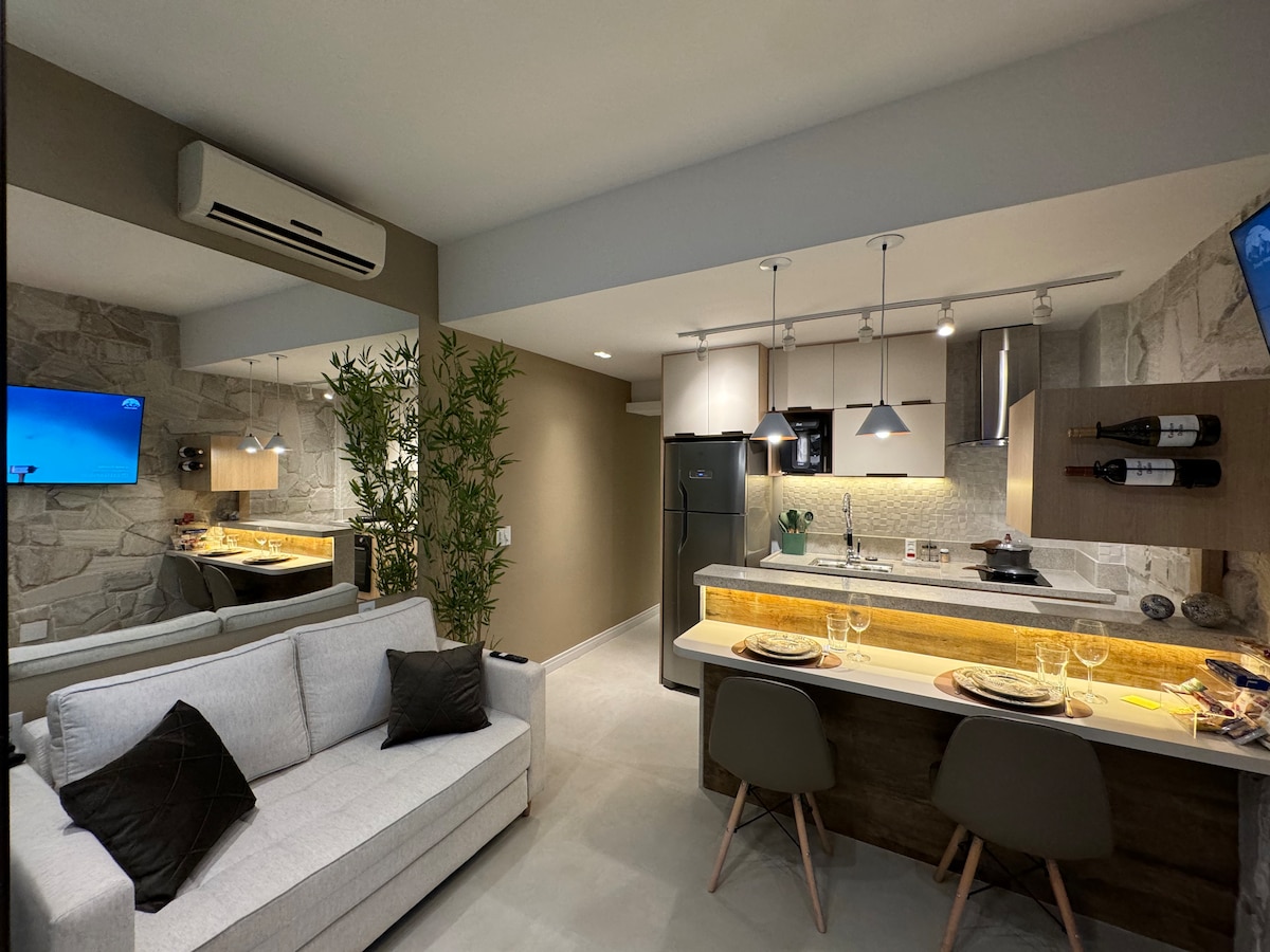 A spacious studio is presented with a modern kitchen area featuring sleek cabinetry and a dining space for two. A cozy sofa with two accent pillows is positioned nearby. Ambient lighting enhances the neutral color palette, creating a harmonious and functional living environment.