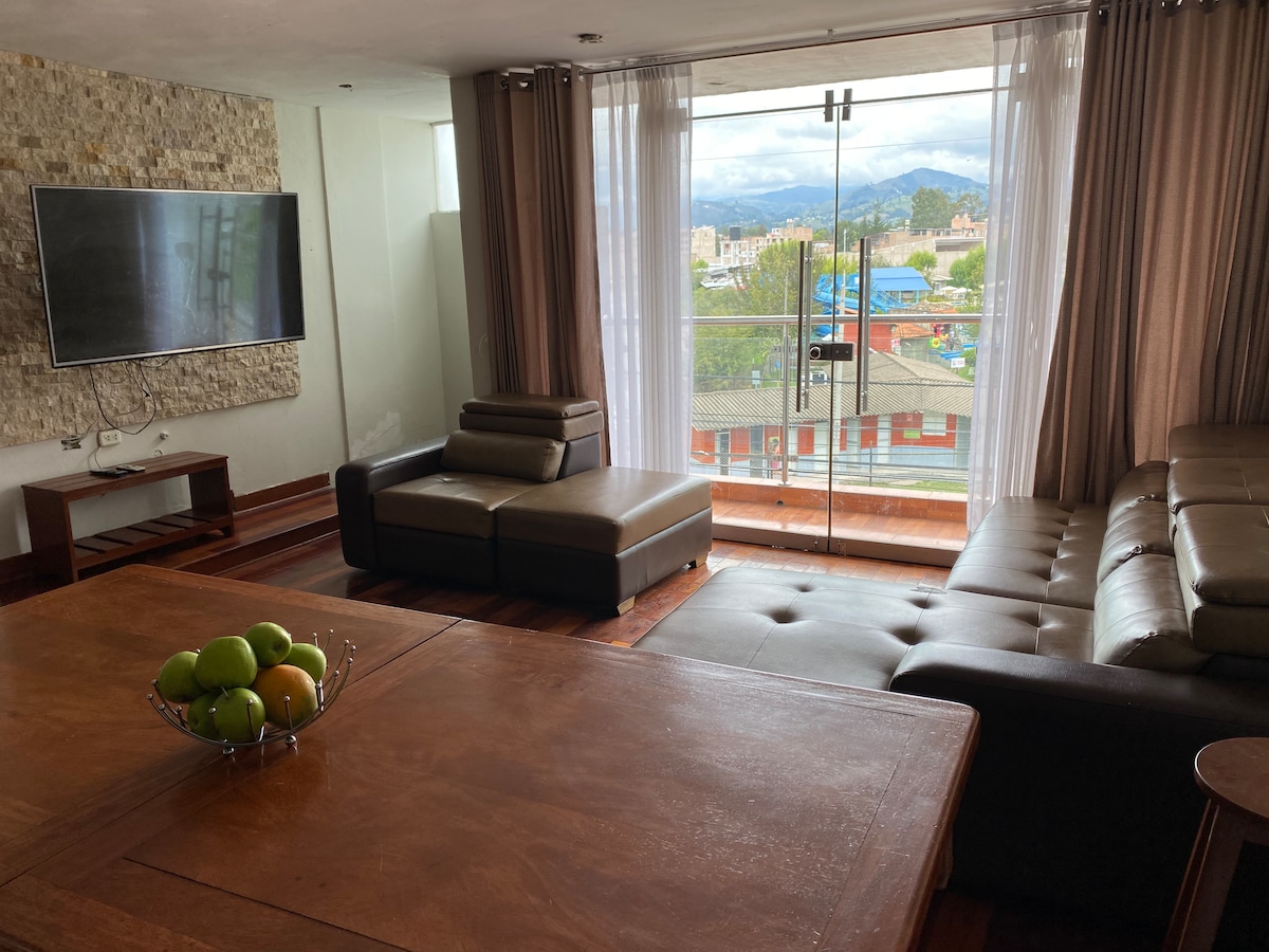 A living area is composed of a spacious sectional sofa and a modern television mounted on a stone-accented wall. Large glass doors open to a balcony showcasing views of the city and surrounding mountains, while natural light brightens the space.