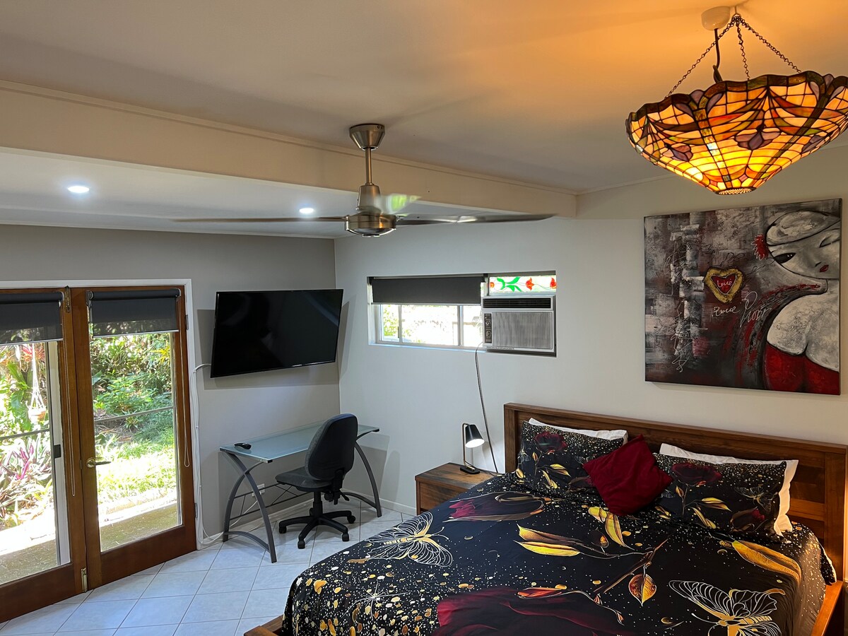A spacious bedroom features a large bed adorned with a colorful bedspread. A ceiling fan provides ventilation, and a desk with a chair is positioned next to a flat-screen TV. Glass doors offer a view of the tropical garden, while natural light filters through the windows.