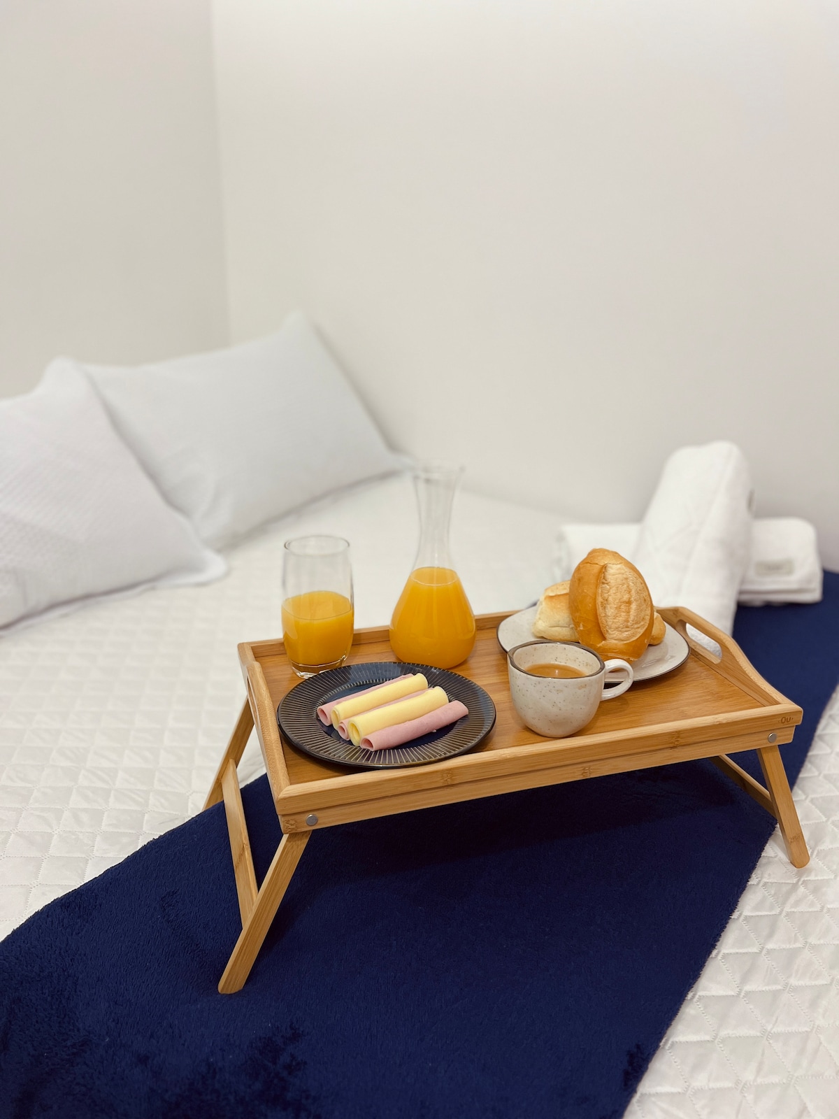 A breakfast tray features two glasses of orange juice, a carafe, and a plate with colorful pastries. A cup of coffee is positioned alongside freshly baked rolls. The bedding is white, with a cozy navy throw displayed across the bed.