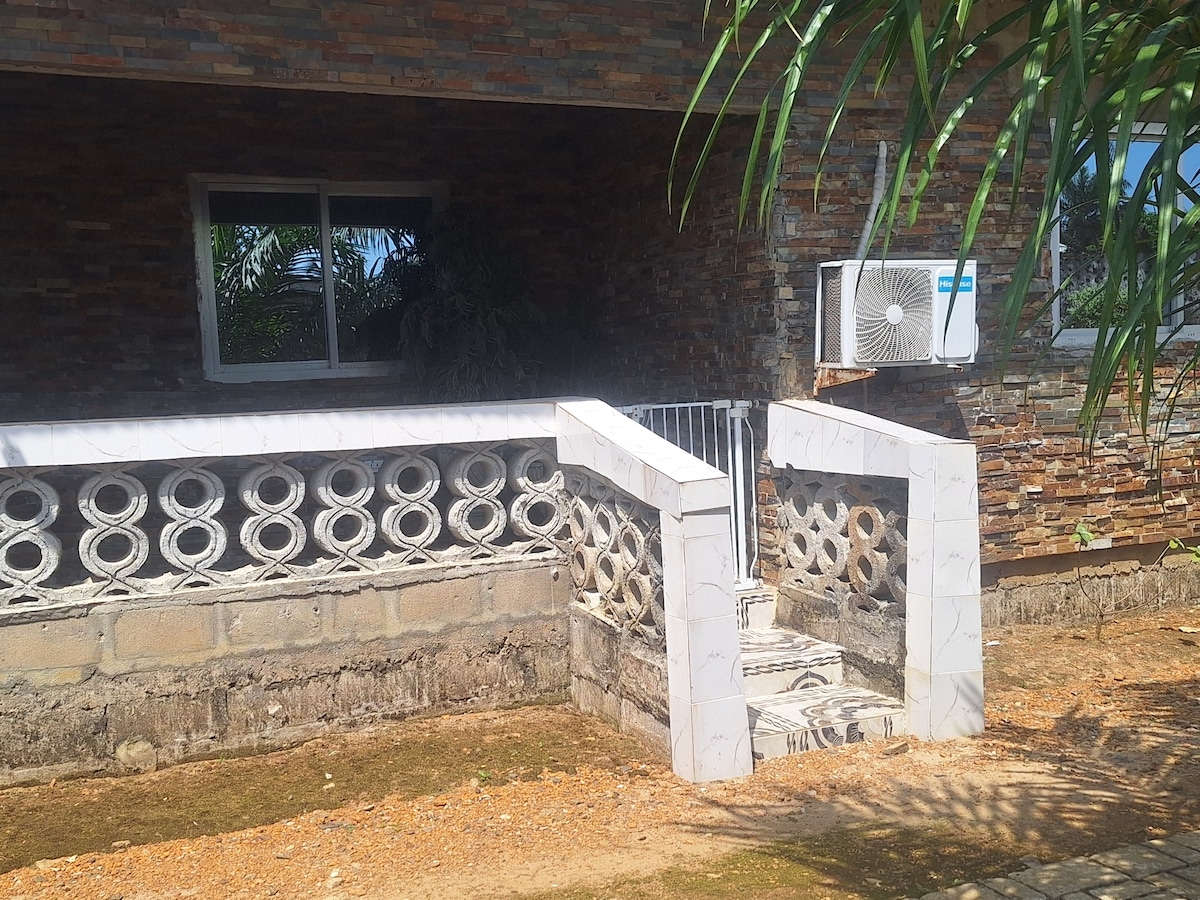 A gated entrance features a unique stone wall design, complemented by a white staircase leading to a spacious area. An air conditioning unit is mounted on the wall beside a large window, with greenery visible in the background.