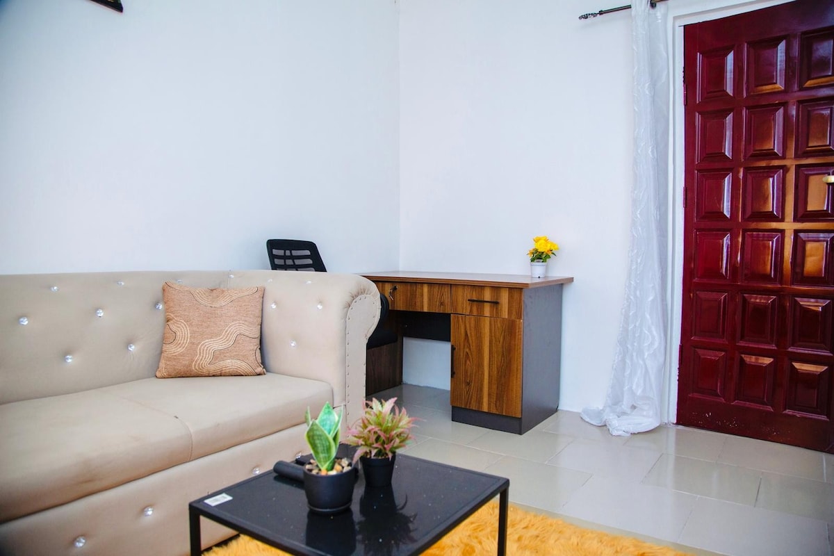 A comfortable lounge area features a light-colored sofa with decorative pillows, complemented by a wooden desk and a small table with potted plants. The backdrop includes a vibrant red door and soft curtain, enhancing a sense of warmth within the space.