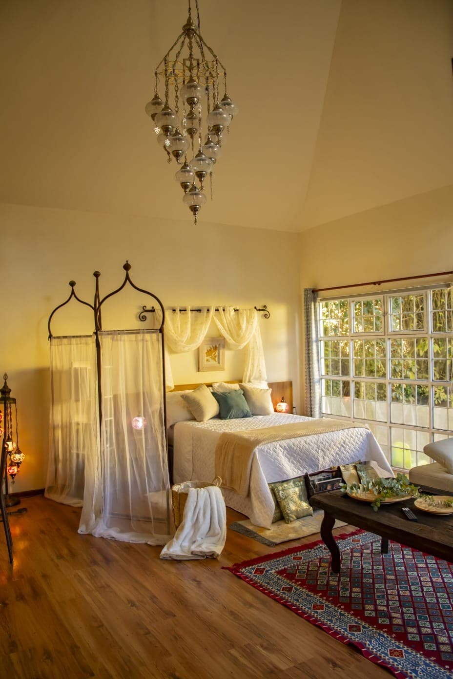 A cozy bedroom is presented with a spacious bed dressed in neutral tones. Soft curtains frame a large window, allowing natural light to illuminate the space. A decorative rug adds texture, while a vintage table complements the setting, adorned with a small display of items.