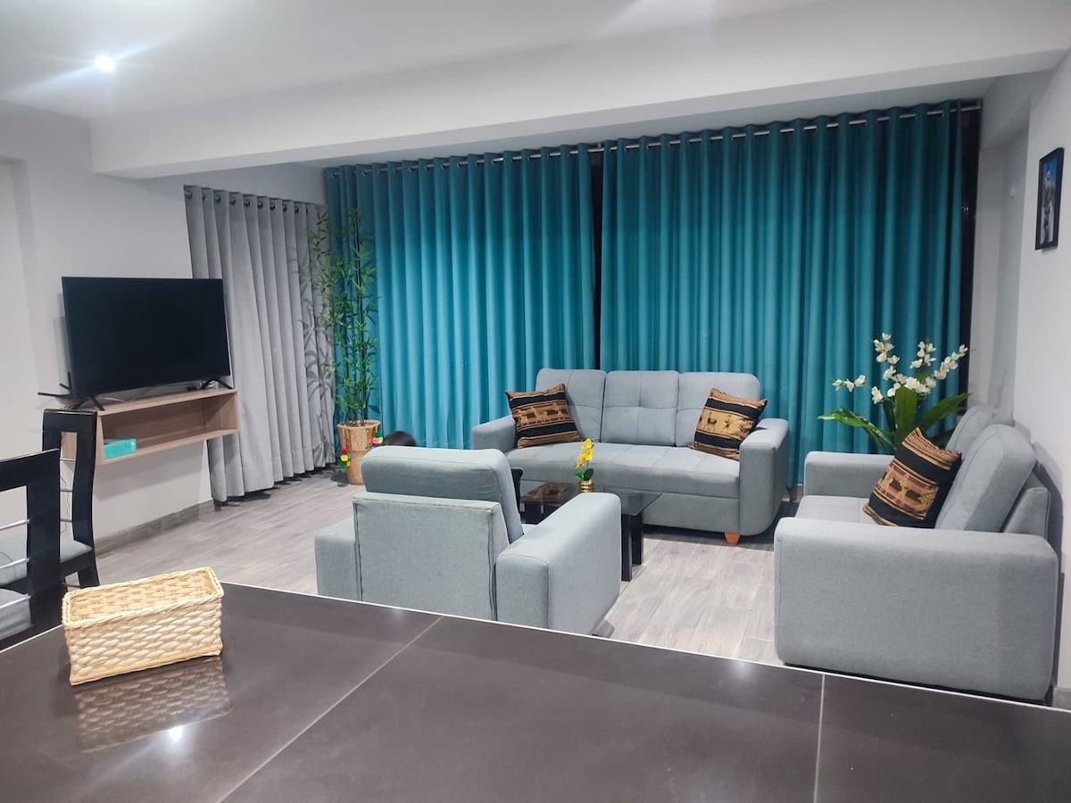 A comfortable living area is presented with multiple soft seating options arranged around a central coffee table. Light blue curtains frame large windows, while a television is mounted on the wall. Decorative plants and a woven basket add a touch of warmth to the ambiance.