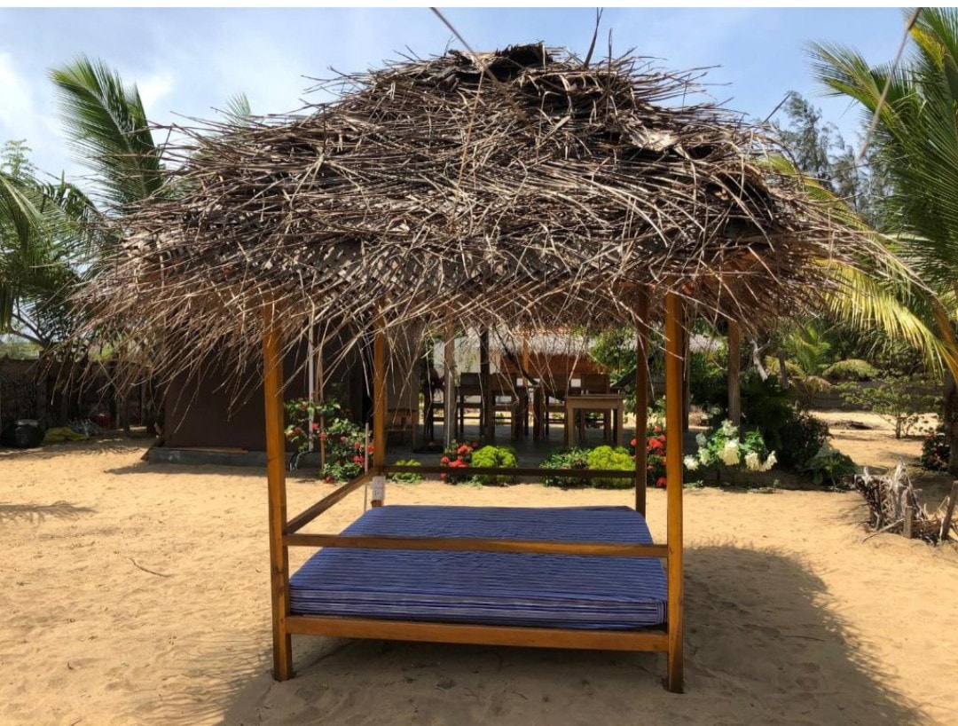A shaded outdoor seating area is presented with a thatched roof supported by a wooden frame, creating a rustic and inviting space. A blue mattress is placed on the sandy ground below, surrounded by vibrant greenery and colorful flowers in the background.