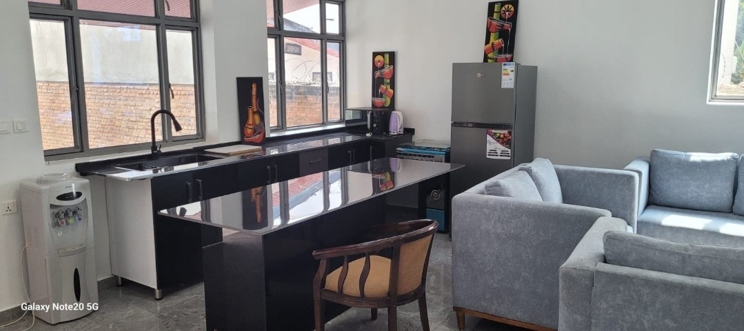A modern kitchen is presented with a sleek black countertop and a matching island. Natural light streams through large windows. An organized refrigerator, visible artwork, and a comfortable seating area with light-colored sofas and an accent chair add to the space.