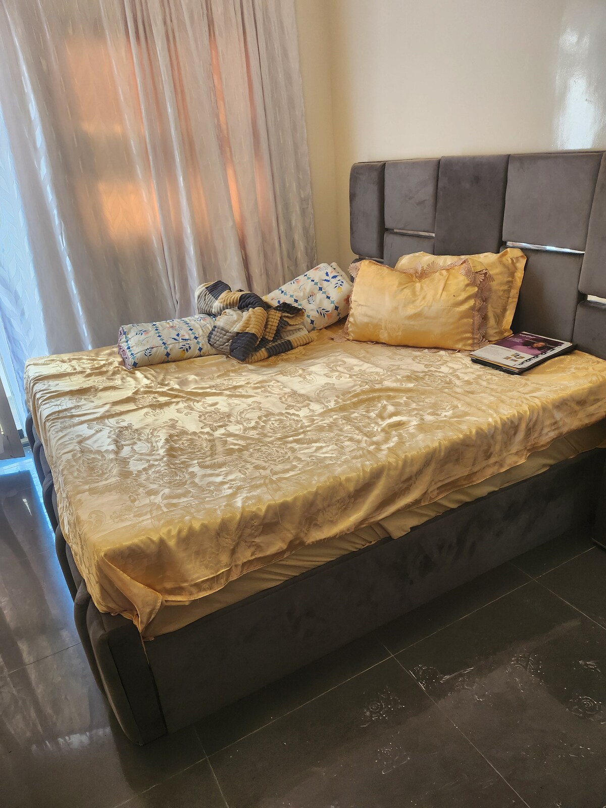 A spacious bed is dressed in soft golden bedding, adorned with decorative pillows. A bedside table holds a magazine, while sheer curtains allow natural light to gently filter into the room. The sleek flooring adds a modern touch to the cozy space.