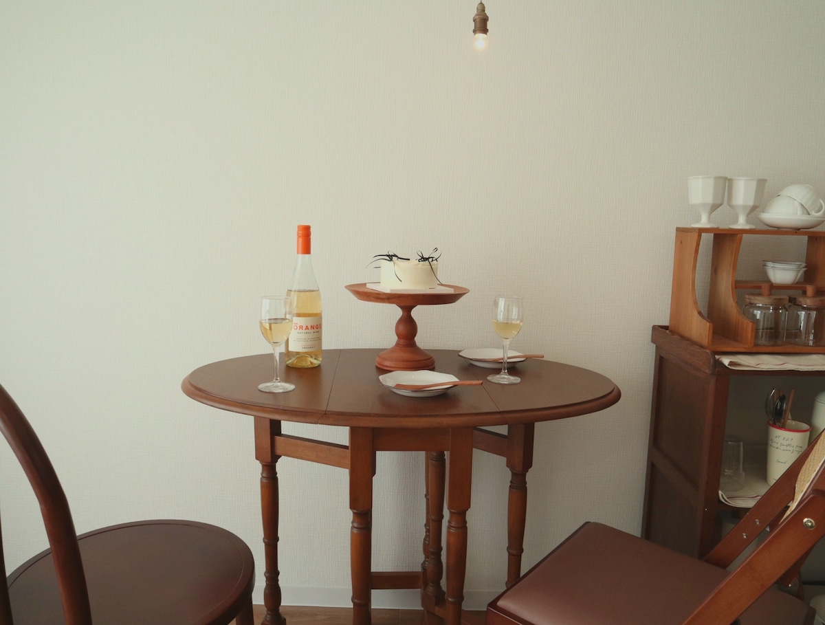 A small wooden dining table is set for two, featuring a bottle of white wine and two glasses. A decorative bowl is placed at the center. The scene is softly illuminated by a pendant light, with wooden chairs positioned around the table.