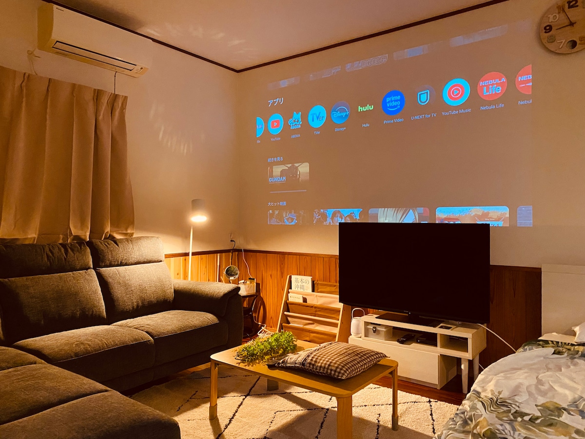 A cozy living area features a large projection screen displaying various apps. A comfortable gray sofa is set adjacent to a modern TV unit. A small coffee table with plants sits in the center, and soft, warm lighting complements the wooden accents.