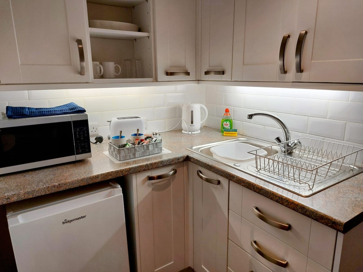 A compact galley kitchen is presented with essential appliances, including a microwave, kettle, and toaster. The counter area features a dish rack and a small sink, with a white tile backsplash enhancing the light. Upper shelves hold cups and additional kitchenware.