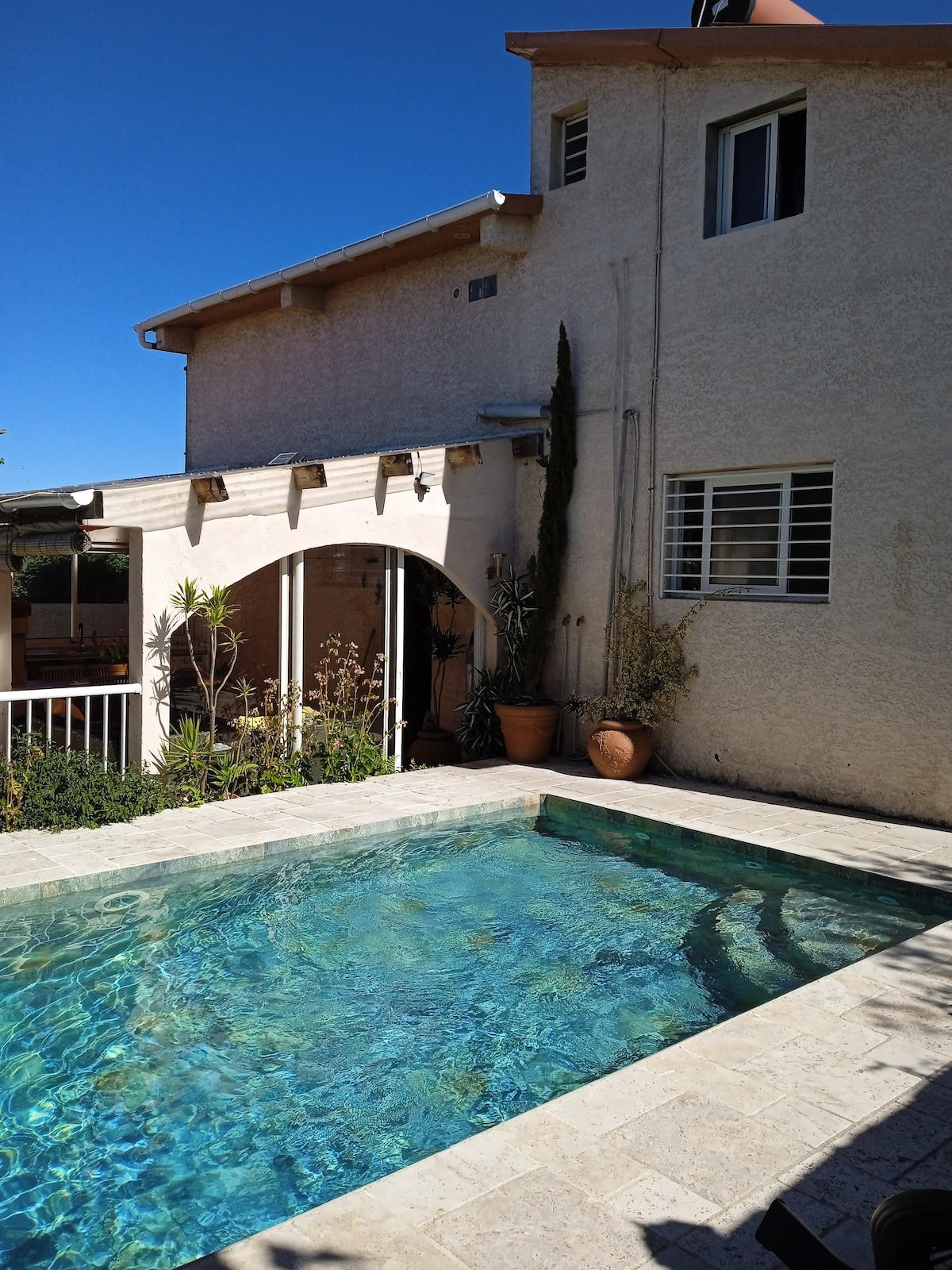 A serene outdoor space features a clear swimming pool surrounded by lush greenery. The pool area is bordered by a natural stone deck and is complemented by potted plants. Sunlight illuminates the inviting water, while the villa's exterior provides a neutral backdrop.