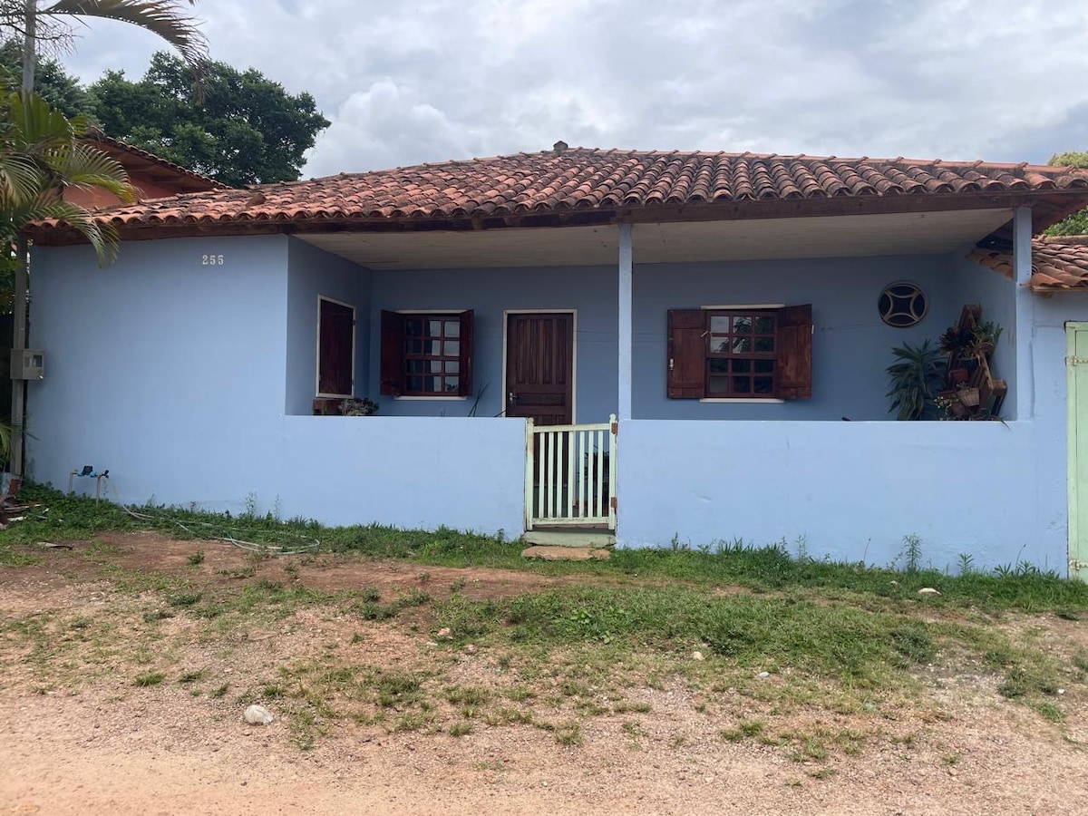The exterior of a light blue house features a traditional tiled roof and wooden shutters. A small porch with a white railing is visible, framed by greenery. The surrounding area includes a gravel pathway leading to the entrance.