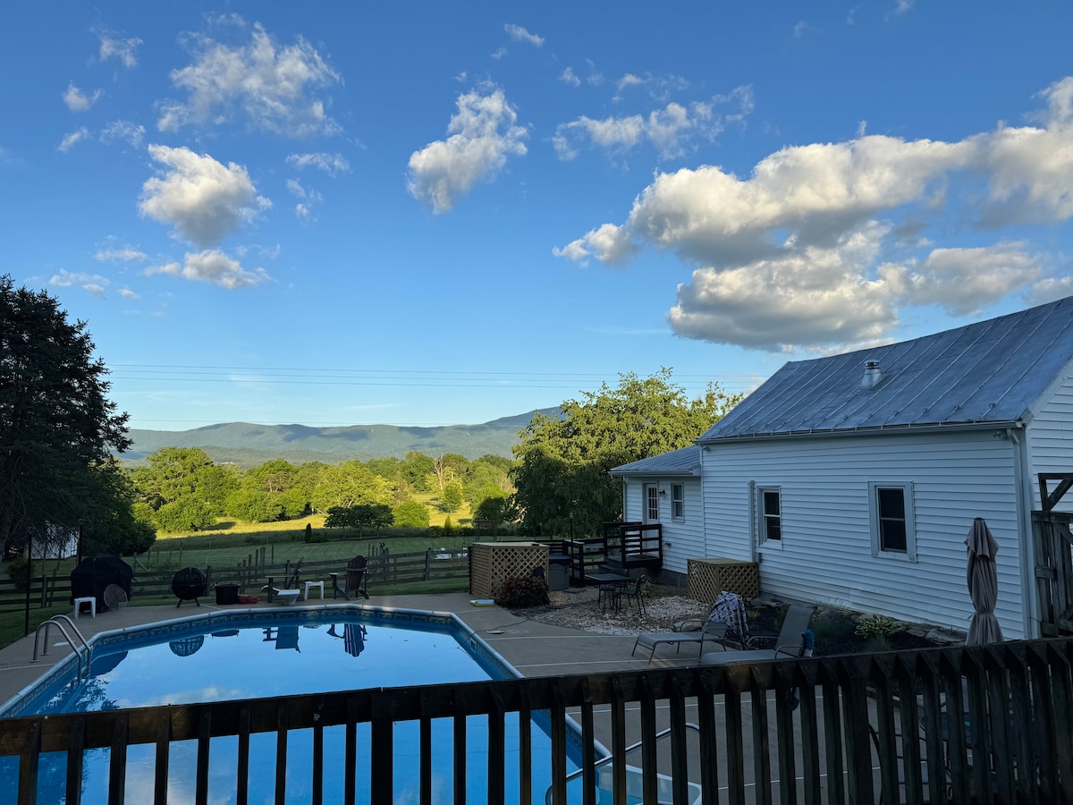 A clear blue sky with fluffy clouds is visible above a private pool, reflecting the surrounding lush landscape. The cottage stands nearby, featuring a white exterior and a metal roof. A fenced area includes relaxing seating options alongside the pool, offering expansive views of the mountains.