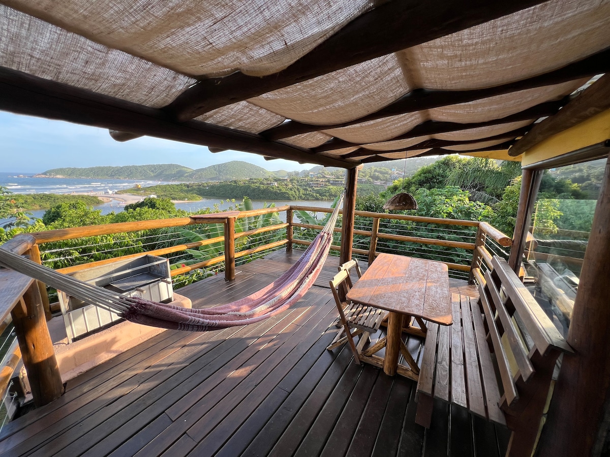 A spacious deck is featured, surrounded by nature and offering views of the mountains and water. A hammock hangs between two supports, and a wooden table with several chairs is positioned for outdoor dining. The deck is covered with a textured canopy, providing shade.