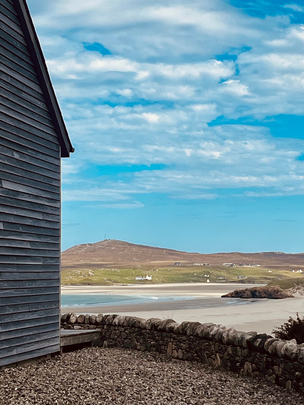 The image depicts the side of a wooden building with horizontal slats, framed by a stone wall. In the background, a sandy shoreline curves towards a distant mountain under a partly cloudy sky, suggesting a serene coastal landscape.