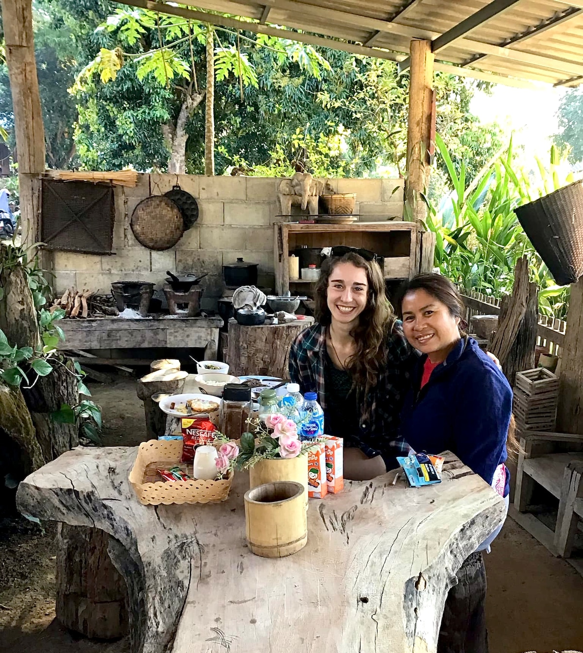 A rustic outdoor kitchen space is showcased, featuring a large wooden table surrounded by various items such as pots, gardening tools, and food supplies. Lush greenery is visible in the background, enhancing the natural ambiance of the setting.