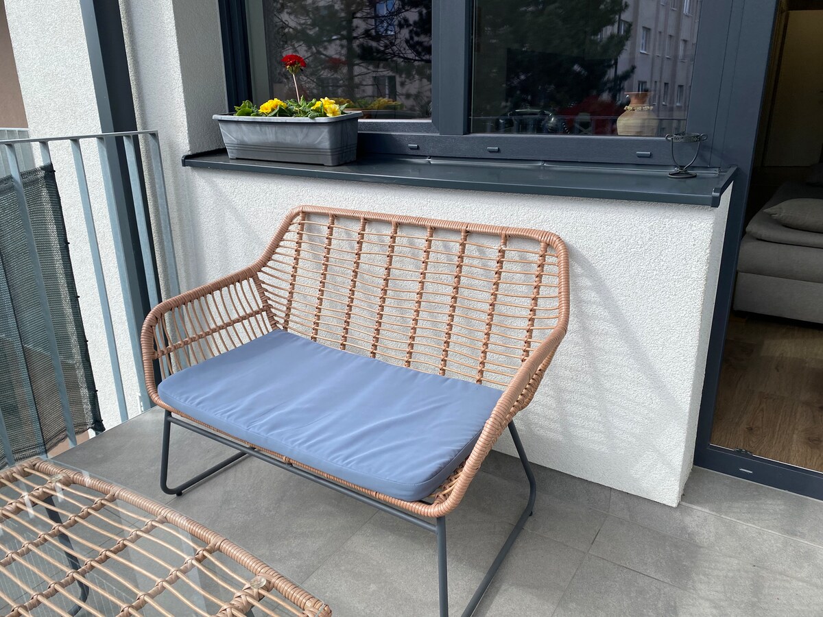 A cozy seating area is presented on the balcony, featuring a two-person rattan bench with a comfortable blue cushion. A planter with colorful flowers is positioned on the windowsill, adding a touch of greenery to the relaxing space.