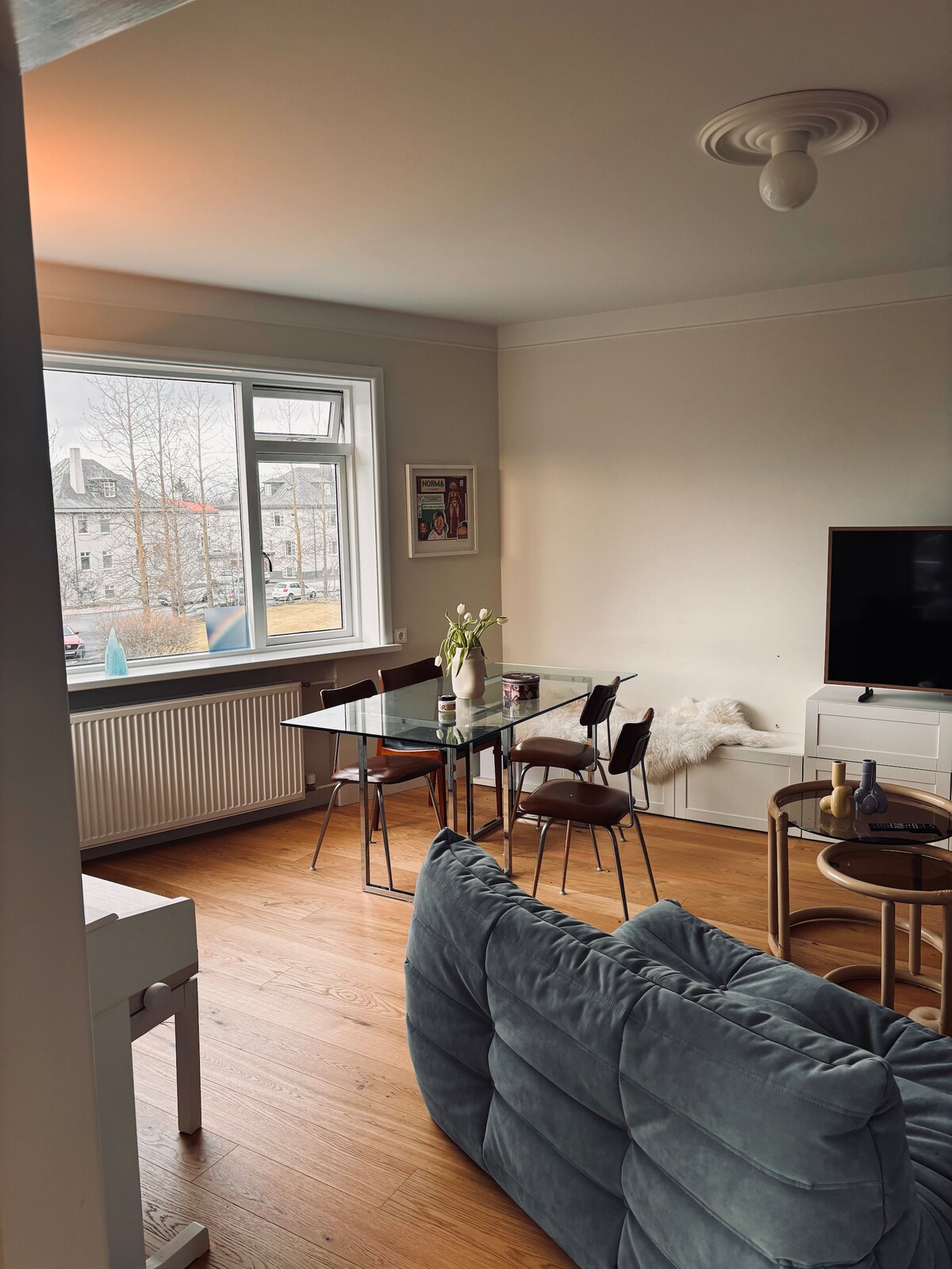 A dining area features a glass table surrounded by four chairs, with a small vase of flowers on top. A soft couch is positioned nearby, complemented by a side table, while a television is placed against the wall, contributing to a cozy living space illuminated by natural light from the window.