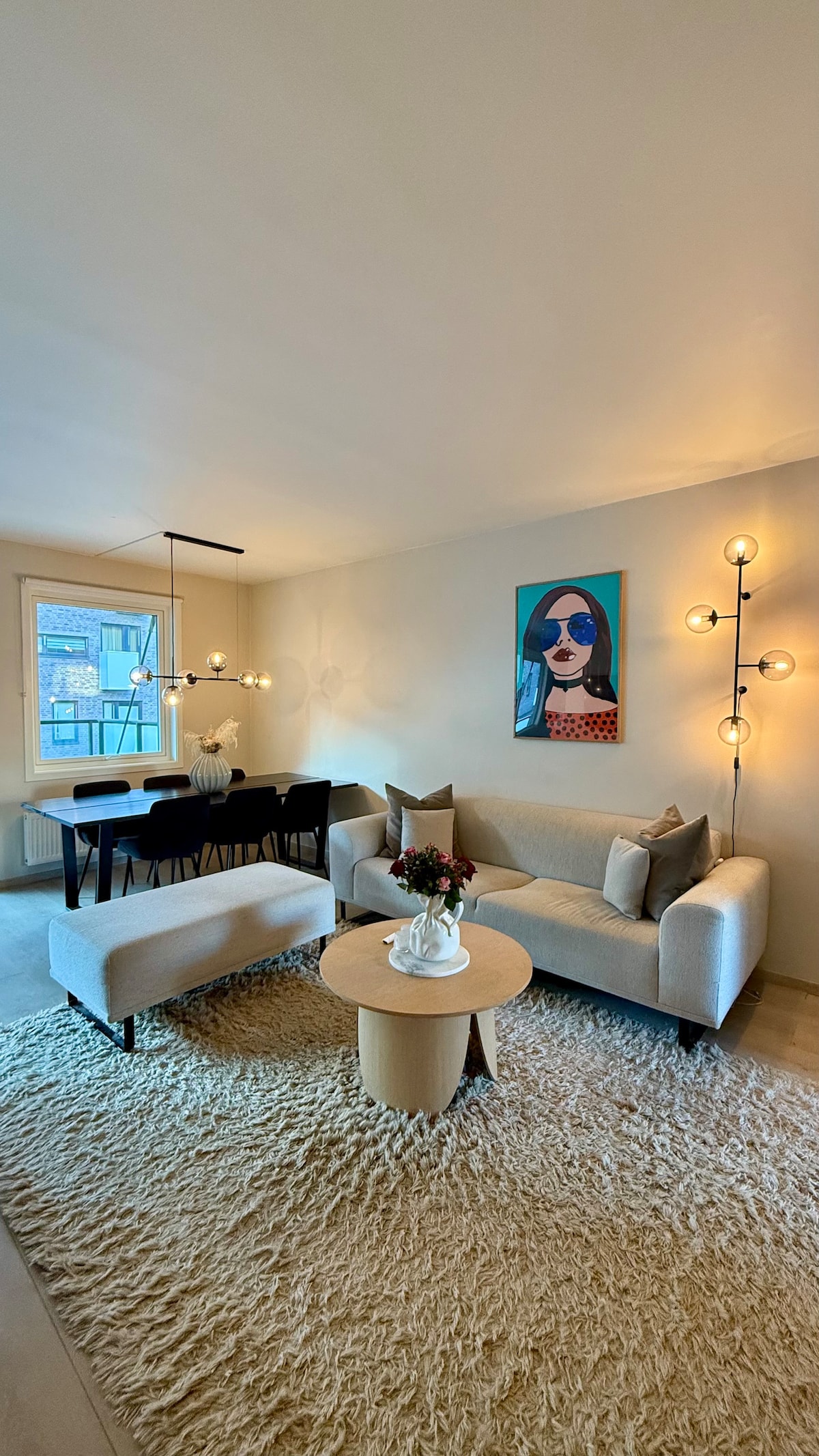 An inviting living area is highlighted by a soft area rug, contemporary seating arrangements, and a circular coffee table. A dining table with chairs is visible in the background, complemented by stylish lighting fixtures. A piece of artwork adds a touch of color to the wall.