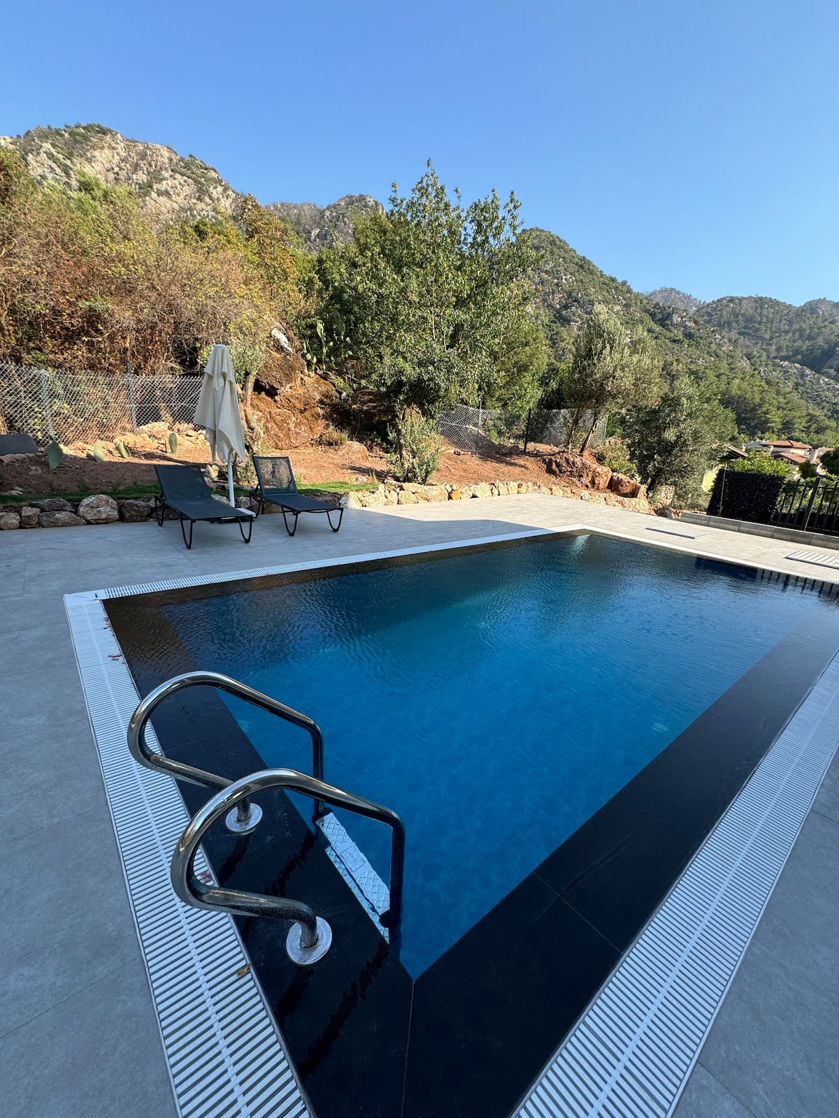 A modern pool is set against a backdrop of lush greenery and distant mountains. Sun loungers are positioned beside the water, and a shaded umbrella offers a relaxing spot. The pool's clean lines and inviting blue surface reflect the clear sky above.