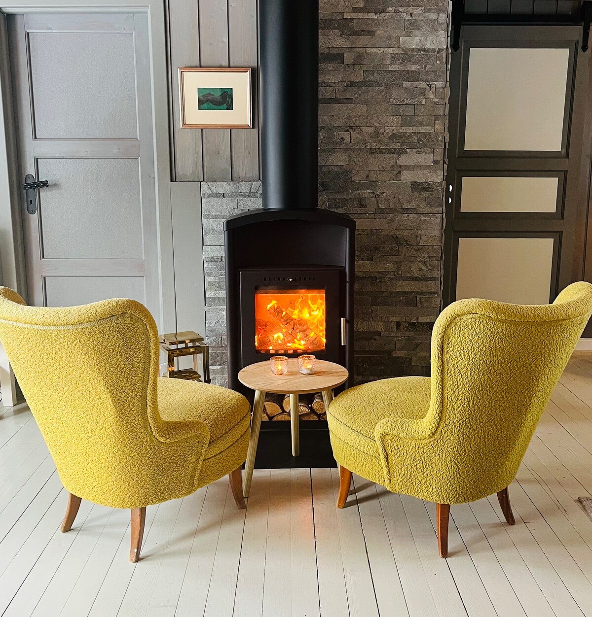 Two yellow upholstered chairs are positioned facing a modern wood stove, creating a cozy conversation area. A small round table sits between the chairs, accompanied by two candle holders. Textured stone walls and light wooden flooring complement the warm ambiance.