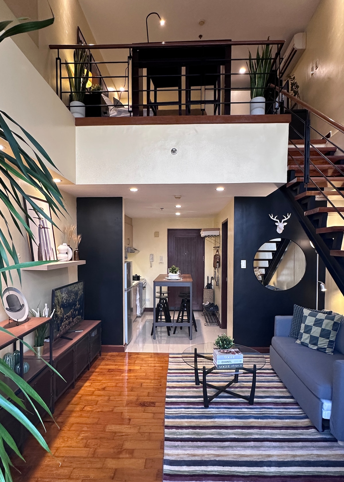 The spacious living area features a comfortable sofa, a patterned rug, and a dining table set for four. A staircase leads to the lofted bedroom above. Large windows allow natural light to flood the space, complemented by decorative plants and modern furnishings.