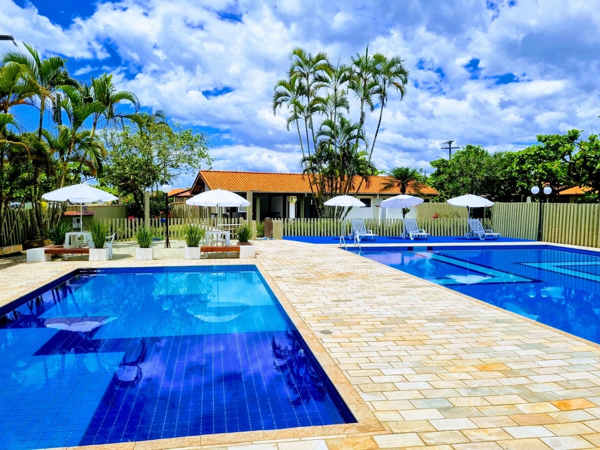 A serene outdoor area features two inviting swimming pools surrounded by beige stone tiles. Sun loungers and umbrellas are placed around the pools, with greenery and palm trees enhancing the tranquil setting under a partly cloudy sky.