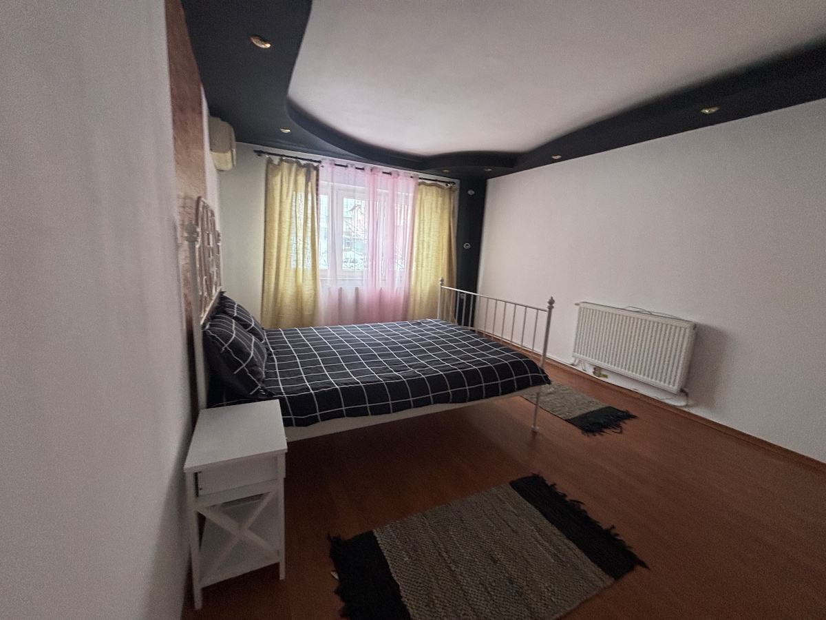 The room features a spacious bed adorned with a checkered black and white blanket. Natural light enters through a window with soft curtains, illuminating the warm wooden flooring. A white nightstand sits adjacent to the bed, complemented by a decorative rug on the floor.