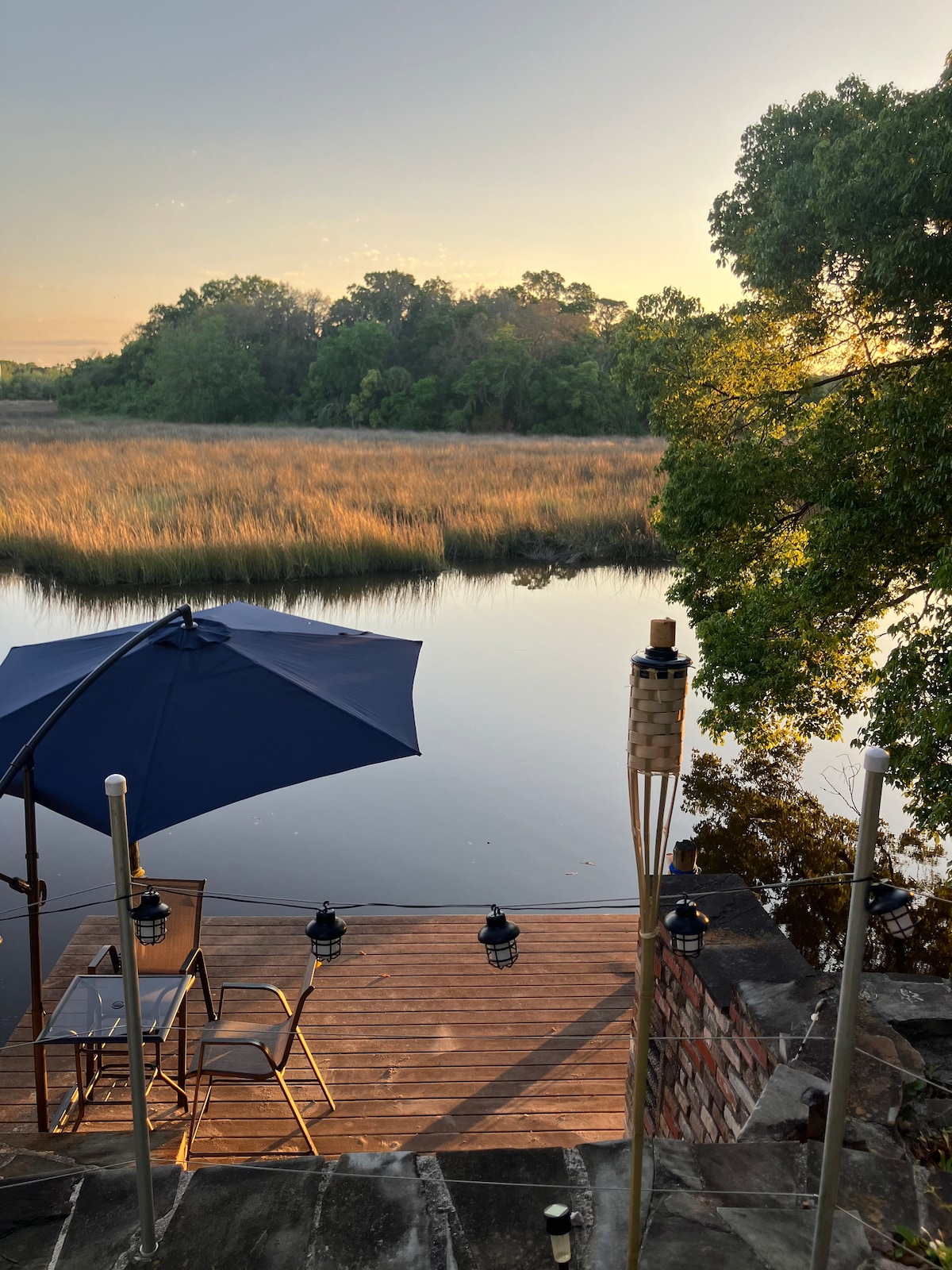 A serene outdoor seating area is shown next to the river, featuring a small wooden deck with a table and two chairs beneath a navy umbrella. Tiki torches line the edge, providing subtle lighting, while the calm water reflects the greenery and golden hues of the setting sun.