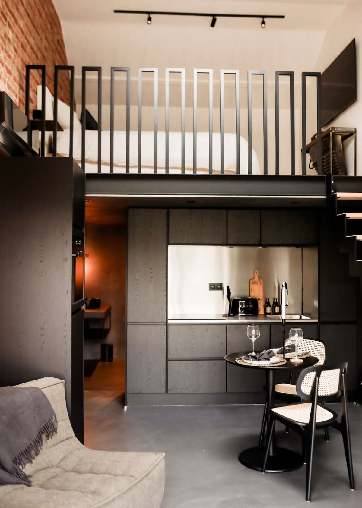 An industrial-style duplex is displayed, featuring a cozy living area with a sofa and a round dining table set for two. A modern kitchen is integrated along one wall. A staircase leads to a lofted sleeping area, with a railing offering a view of the space below.