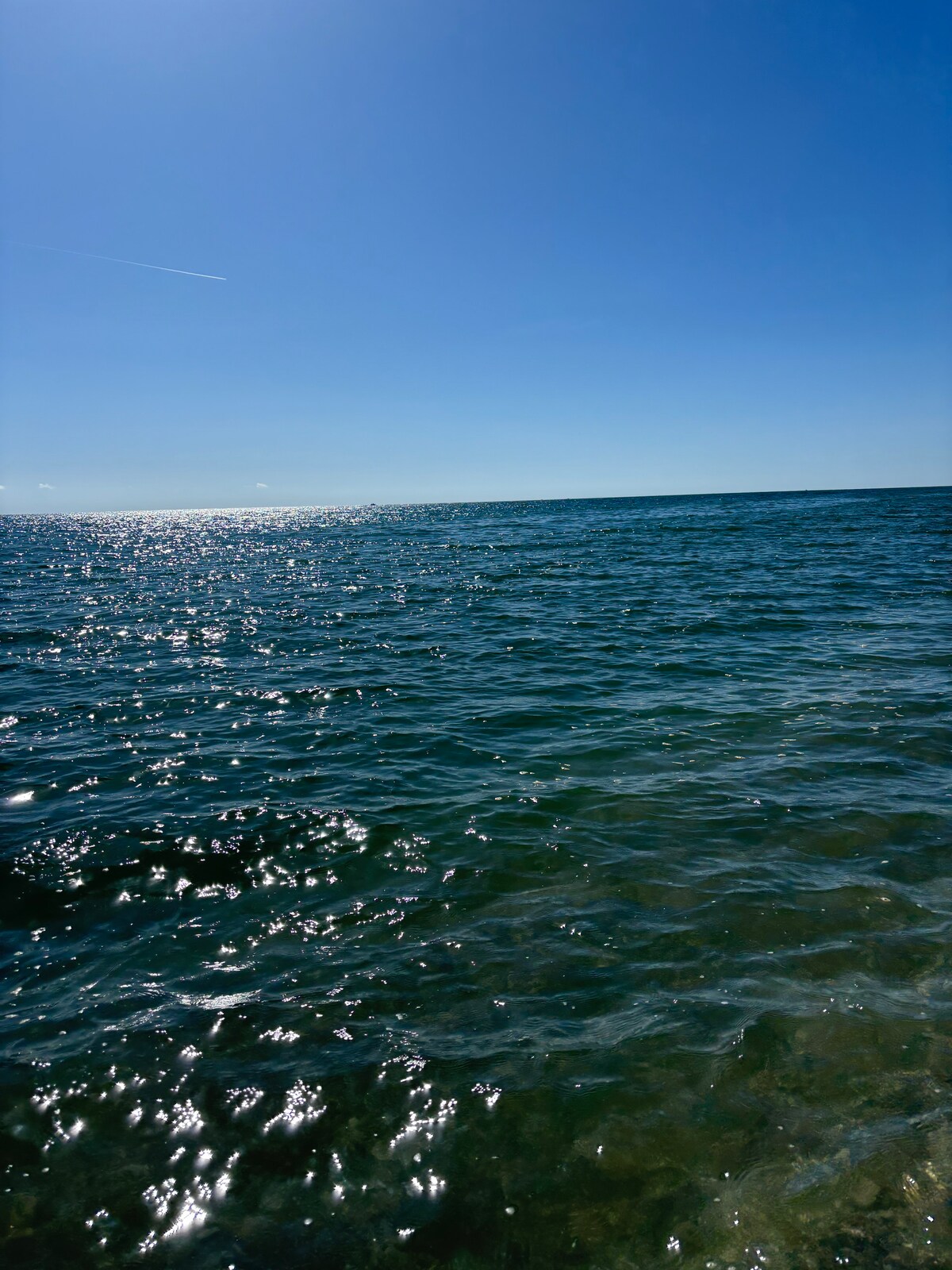 A tranquil view of the sea is captured, displaying calm waters reflecting sunlight. A clear blue sky extends above, providing an unobstructed view of the horizon, while gentle waves create a serene atmosphere.